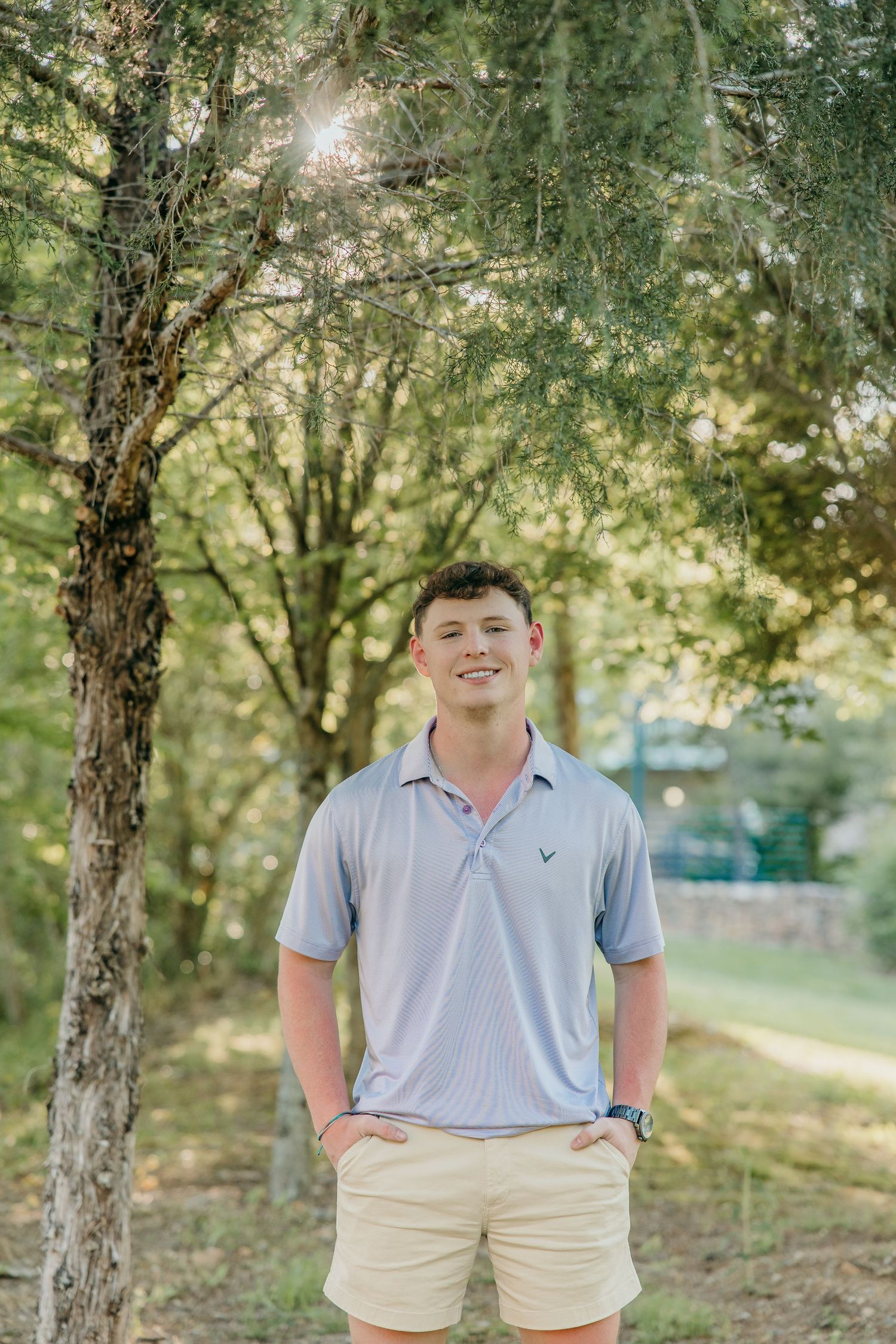 Graduate in polo shirt and shorts poses in wooded area with sunlight filtering through trees.