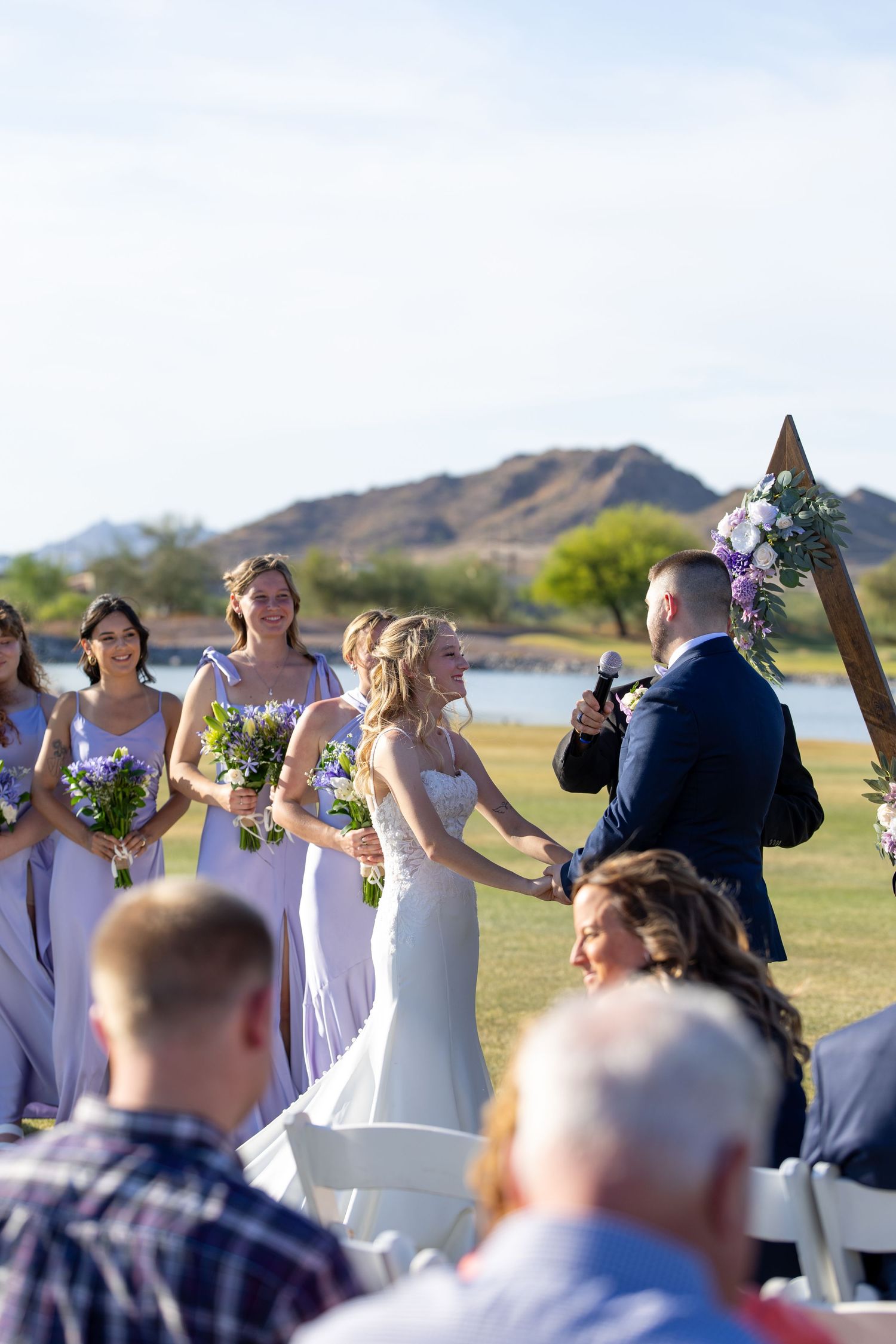 Outdoor wedding ceremony with bride and groom holding hands at wooden arch, bridesmaids in lavender dresses, mountain lake backdrop.