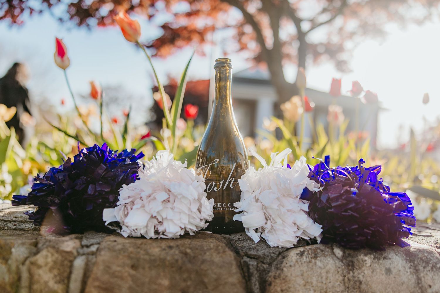 Purple and white flowers with champagne bottle arranged on stone wall during sunset.