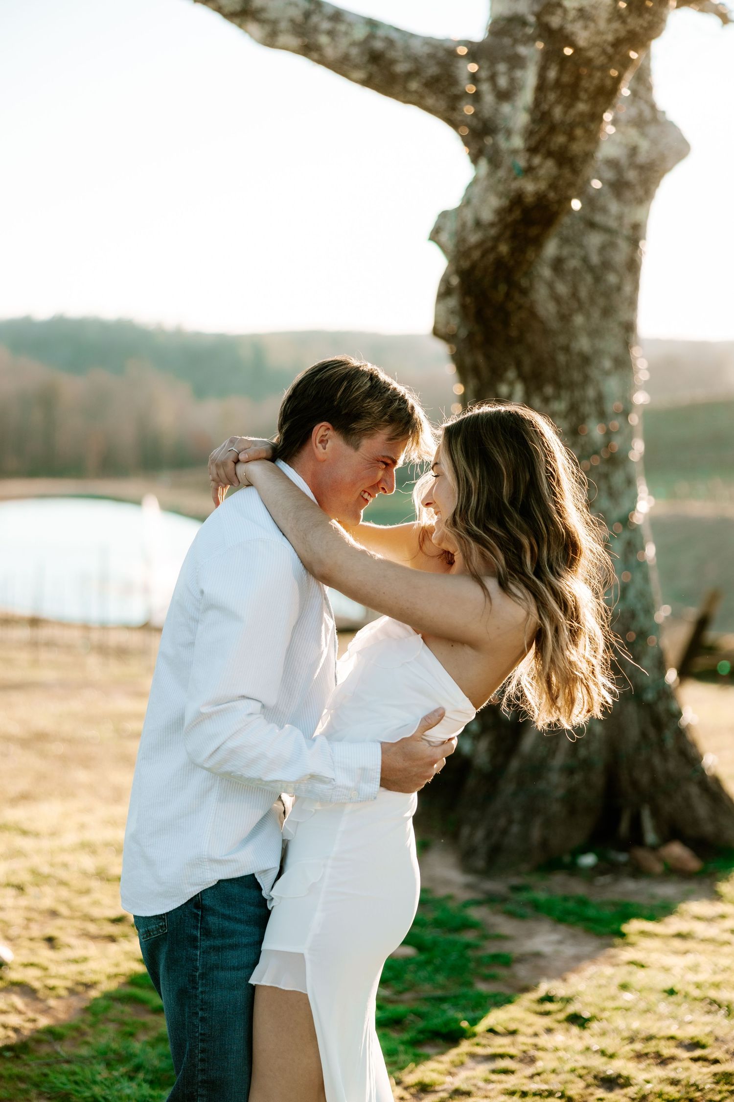 Couple embraces in sunset light beneath large tree during romantic photoshoot.