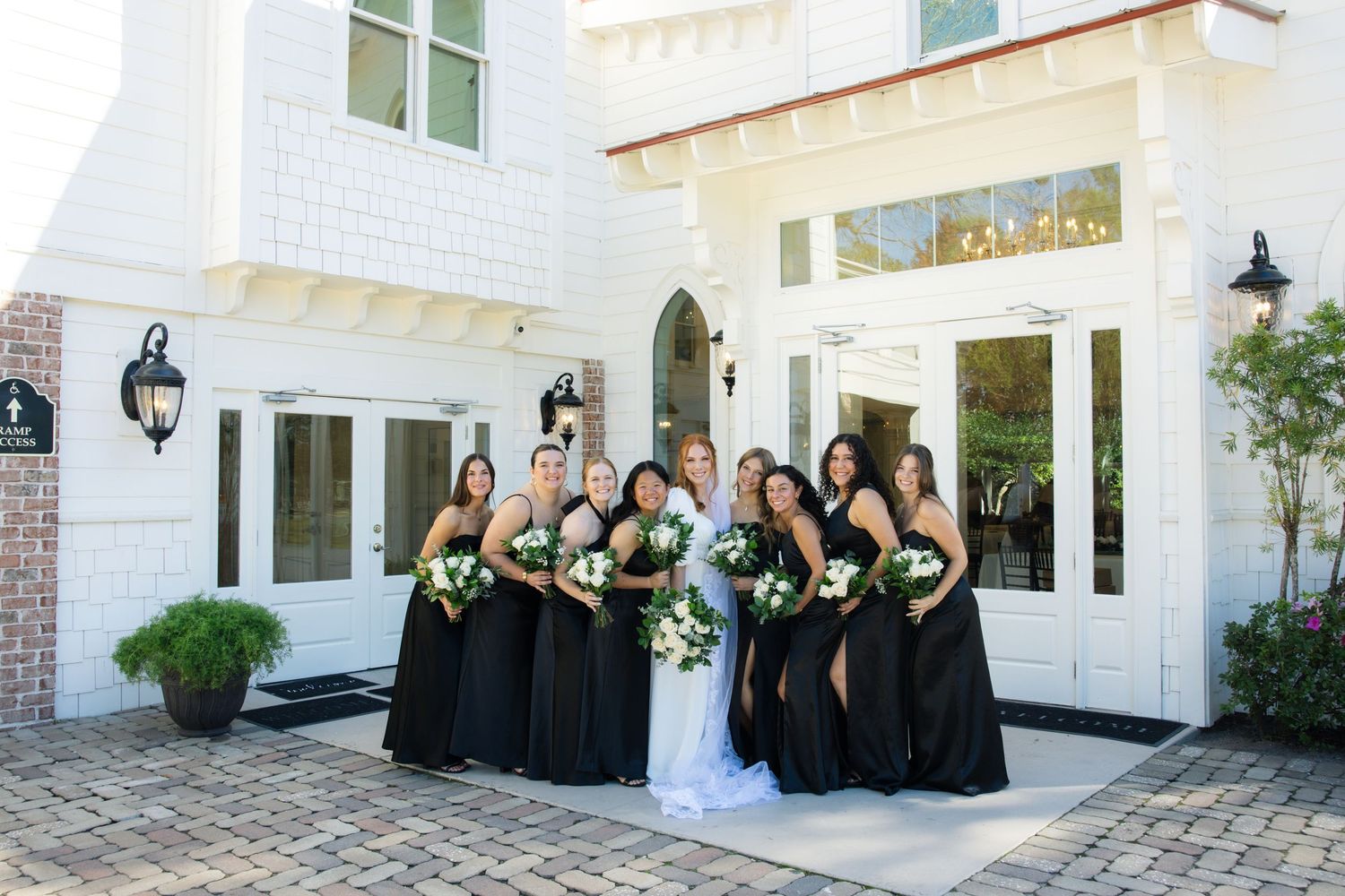 Bride in white gown poses with bridesmaids in black dresses holding bouquets outside elegant white venue.