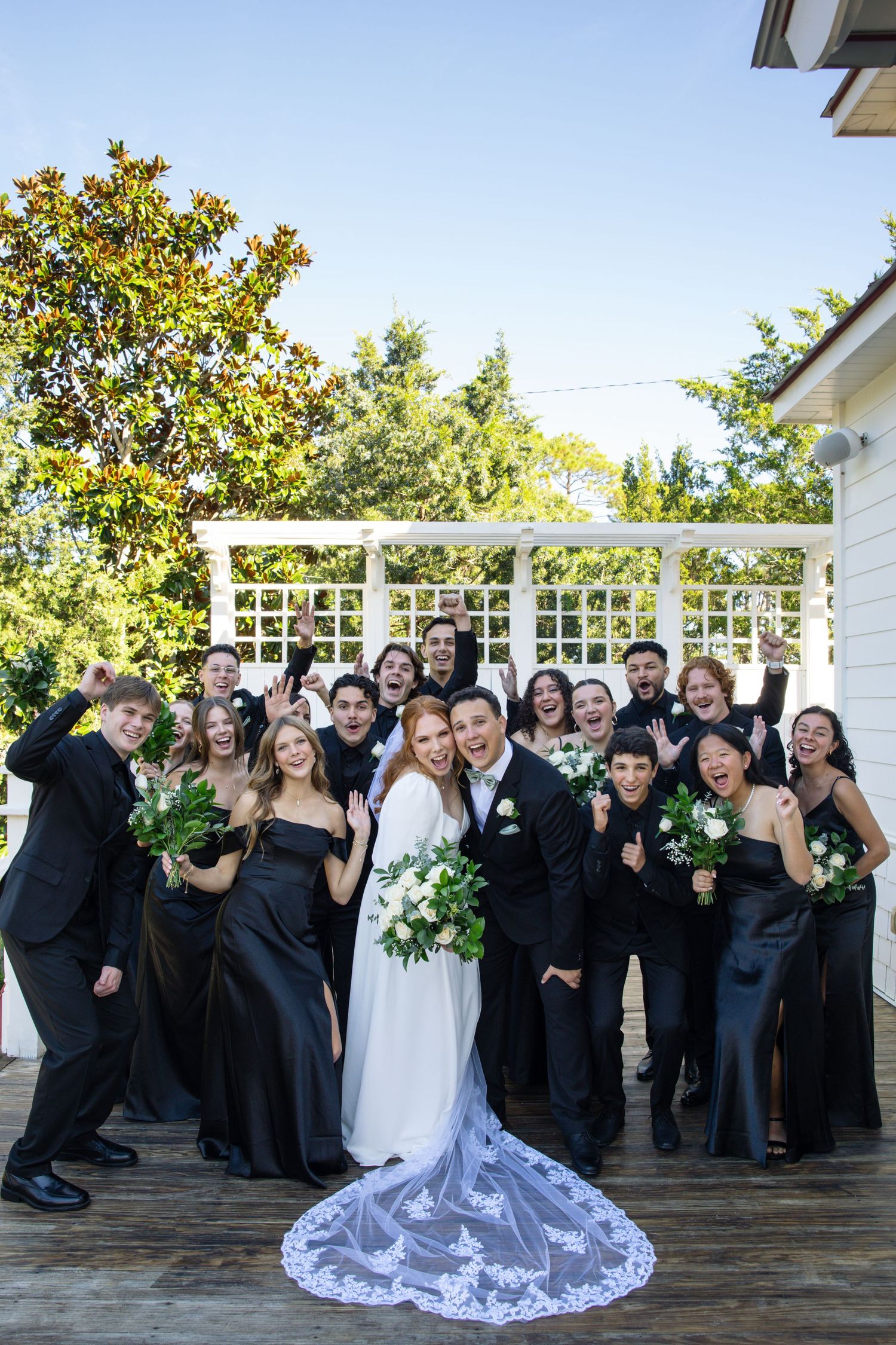 Wedding party poses for fun group photos on wooden deck wearing black and white formal attire.
