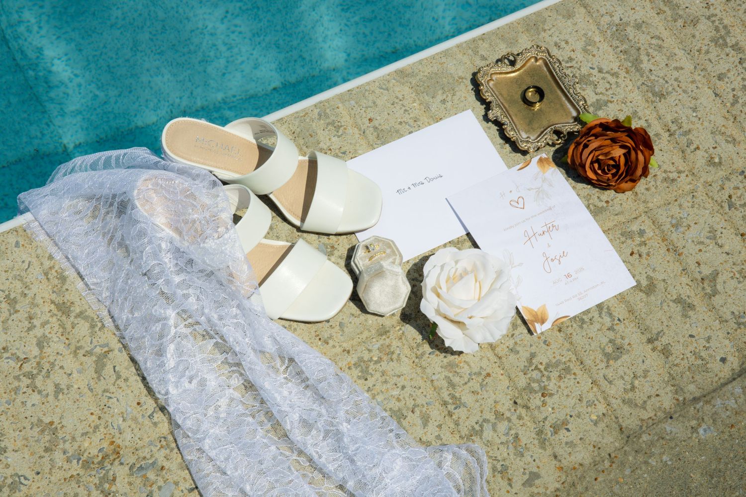 Wedding accessories including white shoes, handwritten notes, lace fabric, and floral decorations arranged on a textured surface.