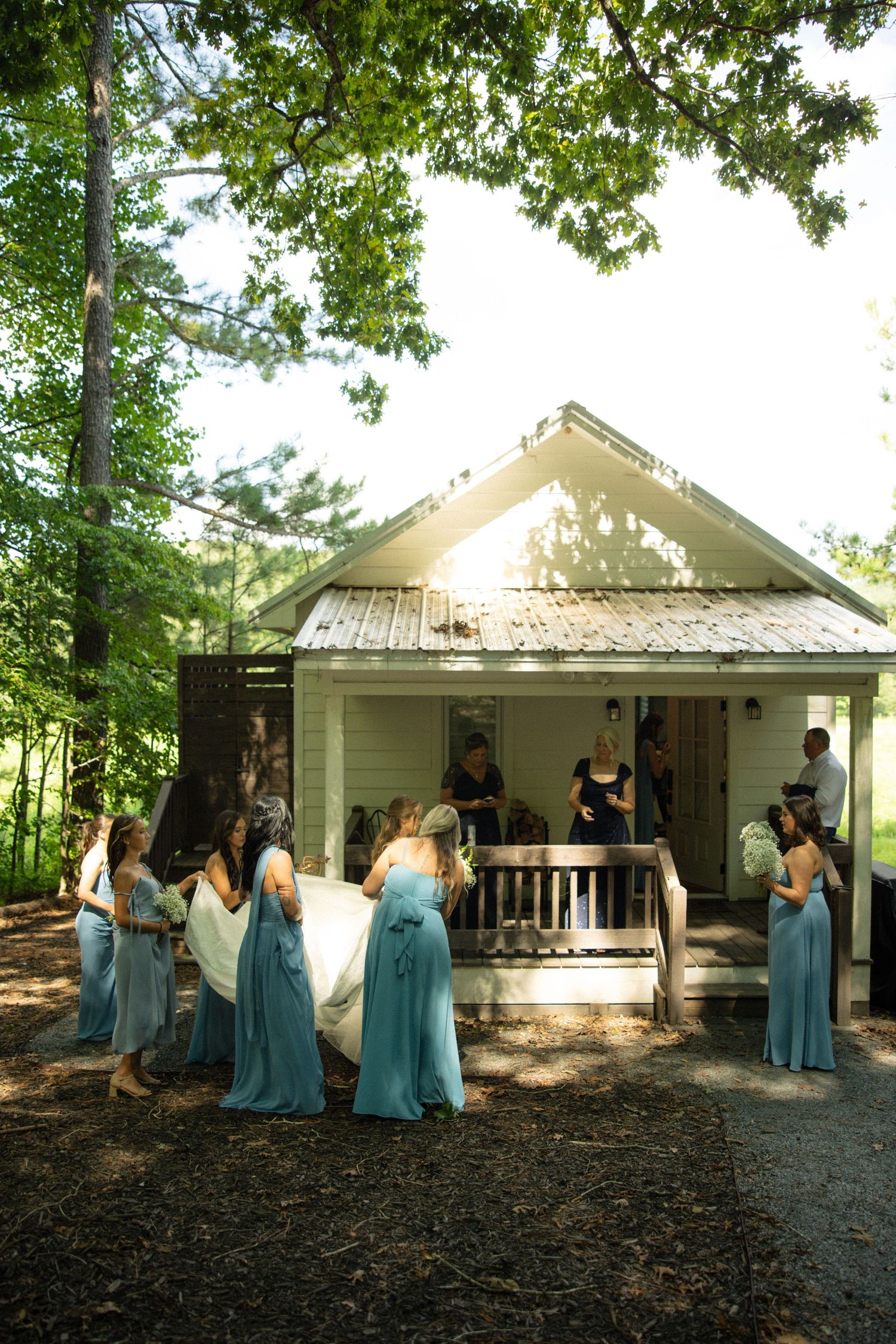 Wedding party members in blue dresses gather outside a rustic white building during an outdoor ceremony.