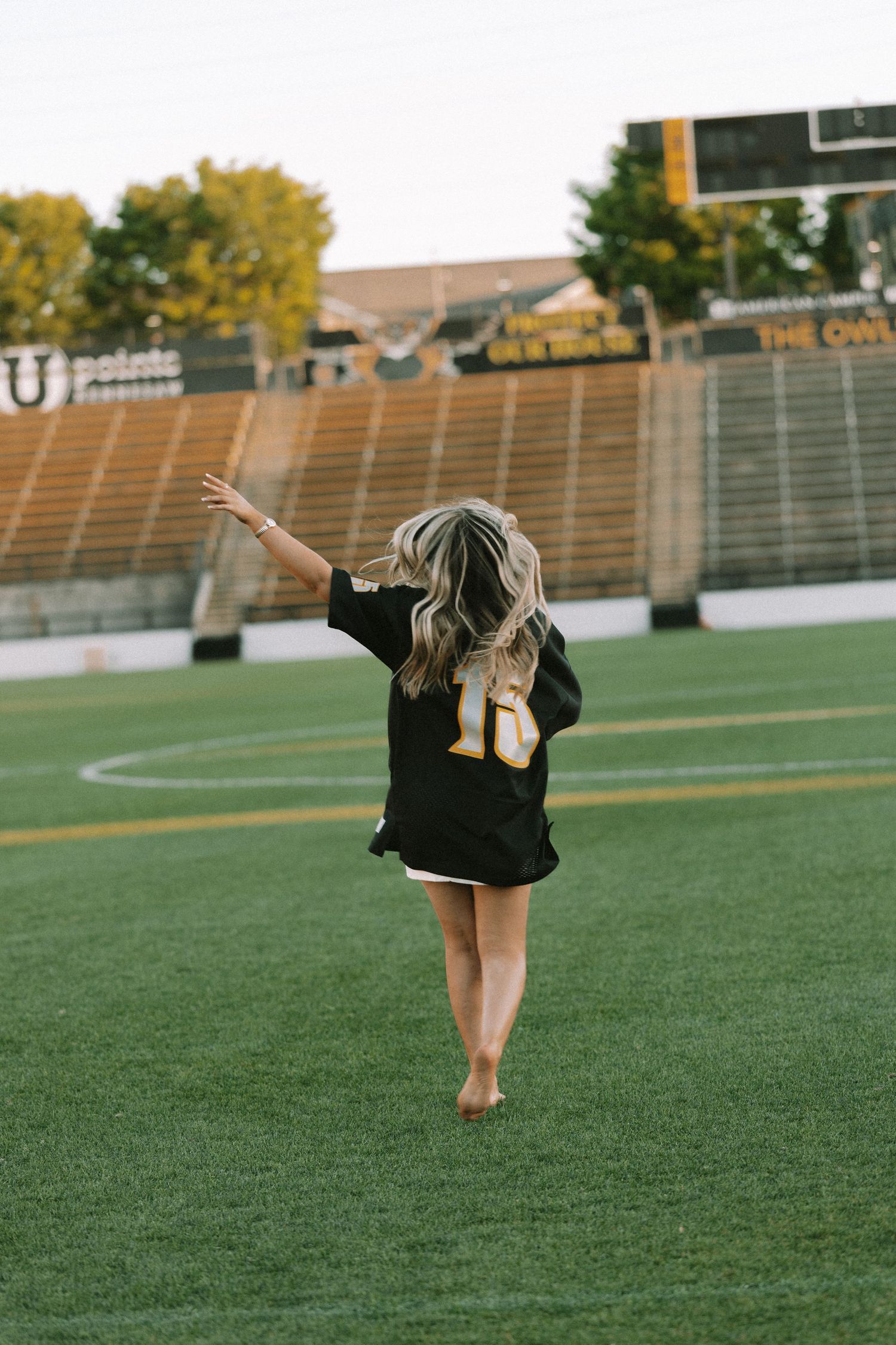 Person in jersey walks barefoot on football field during sunset graduation photoshoot.