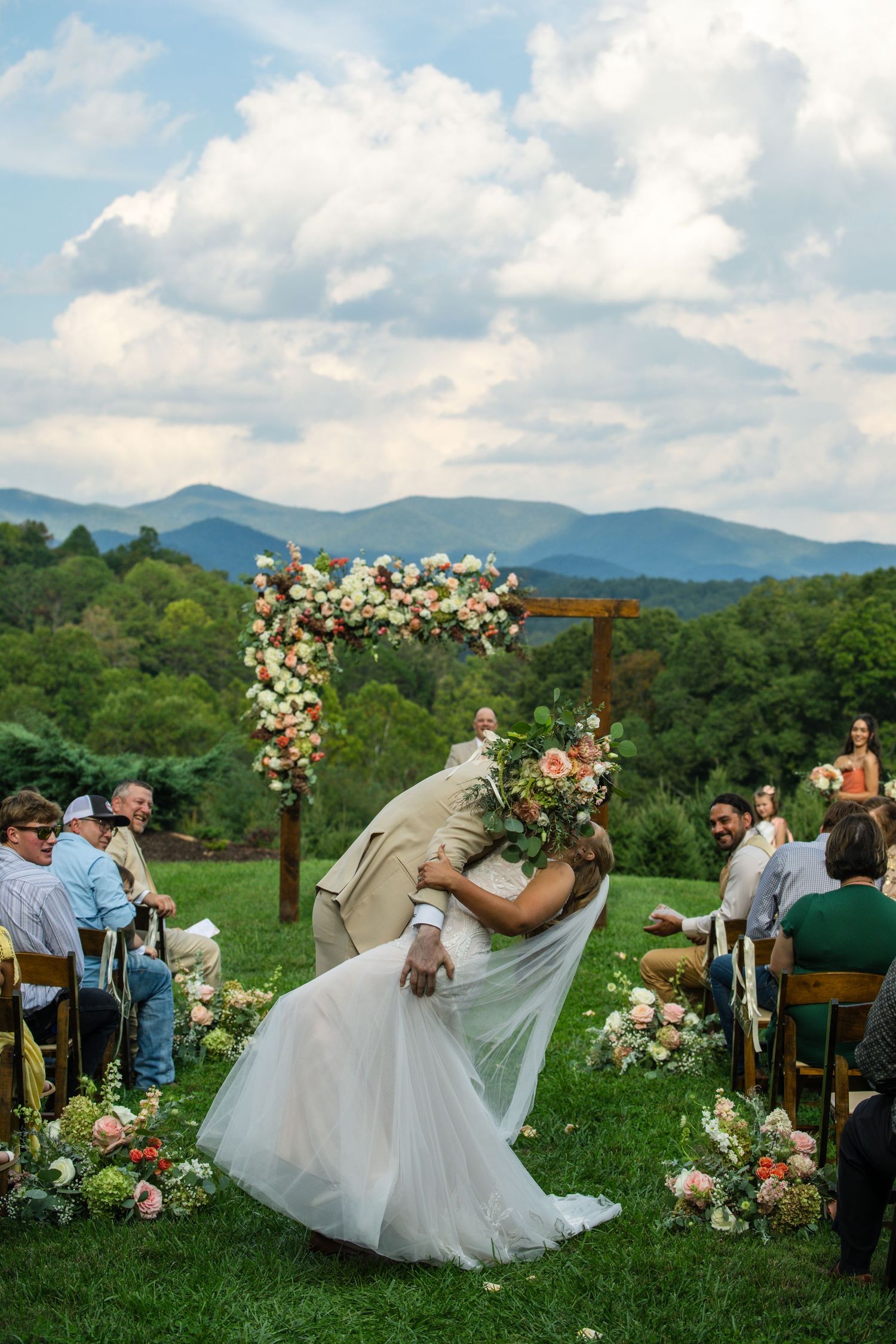Groom dips bride for a kiss at outdoor wedding ceremony with floral arch and mountain backdrop.