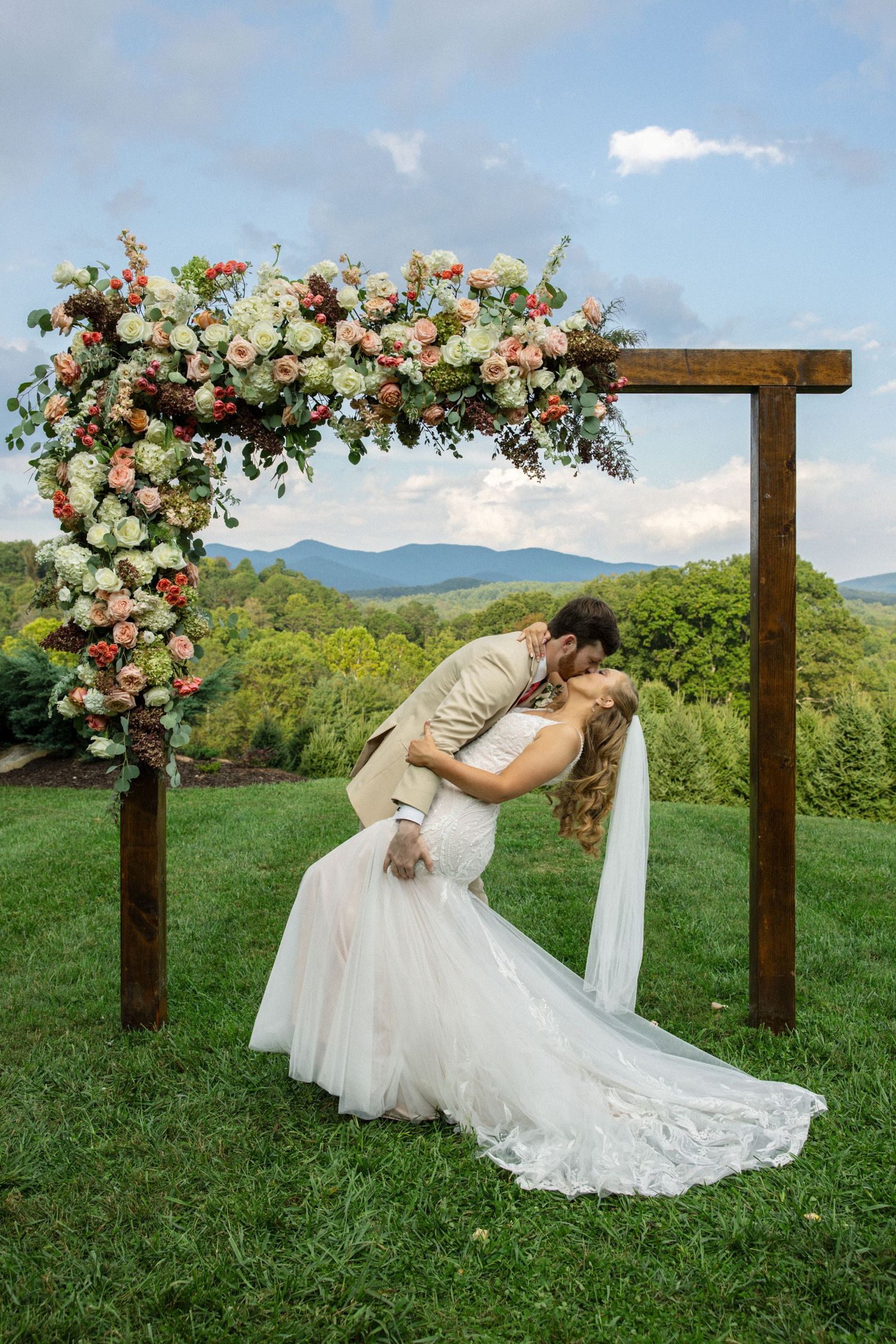 A wedding dress flows gracefully on a grassy hillside beneath a wooden arch adorned with pink and peach roses.
