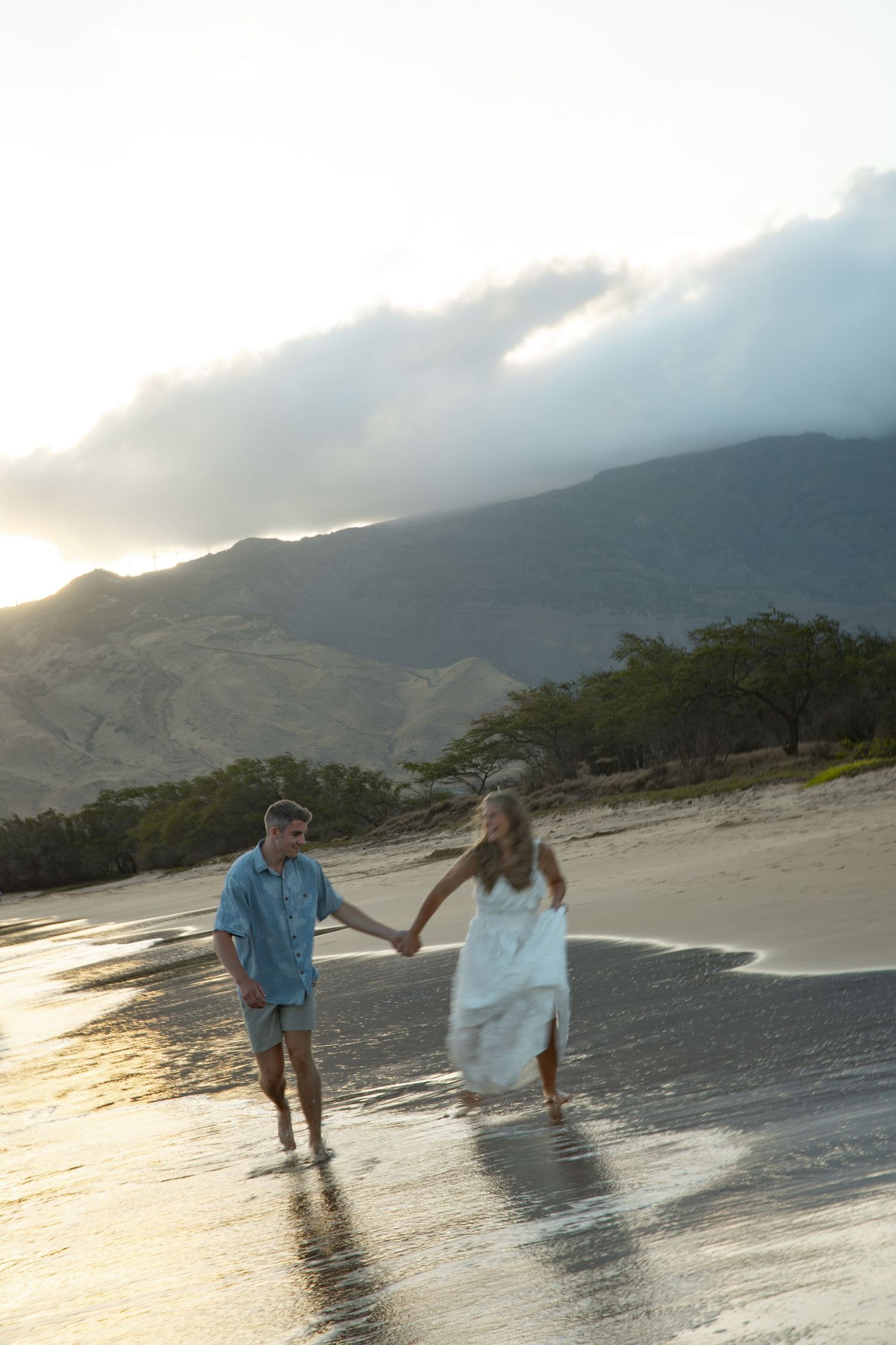 A couple walks along a misty beach at sunset with mountains in the background creating a romantic silhouette.