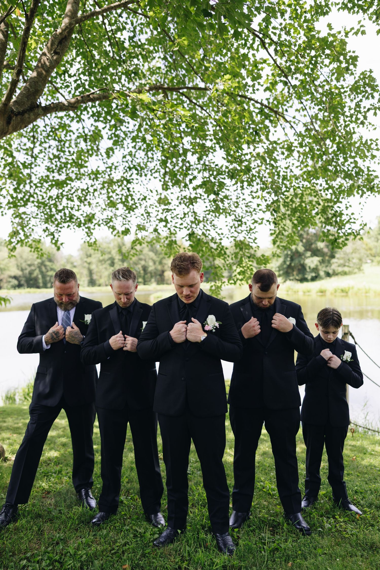 A line of groomsmen in black suits bow their heads while standing under a tree outdoors.