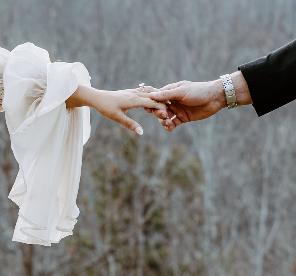 Two hands reaching for each other in an intimate moment, with a flowing white dress in frame.