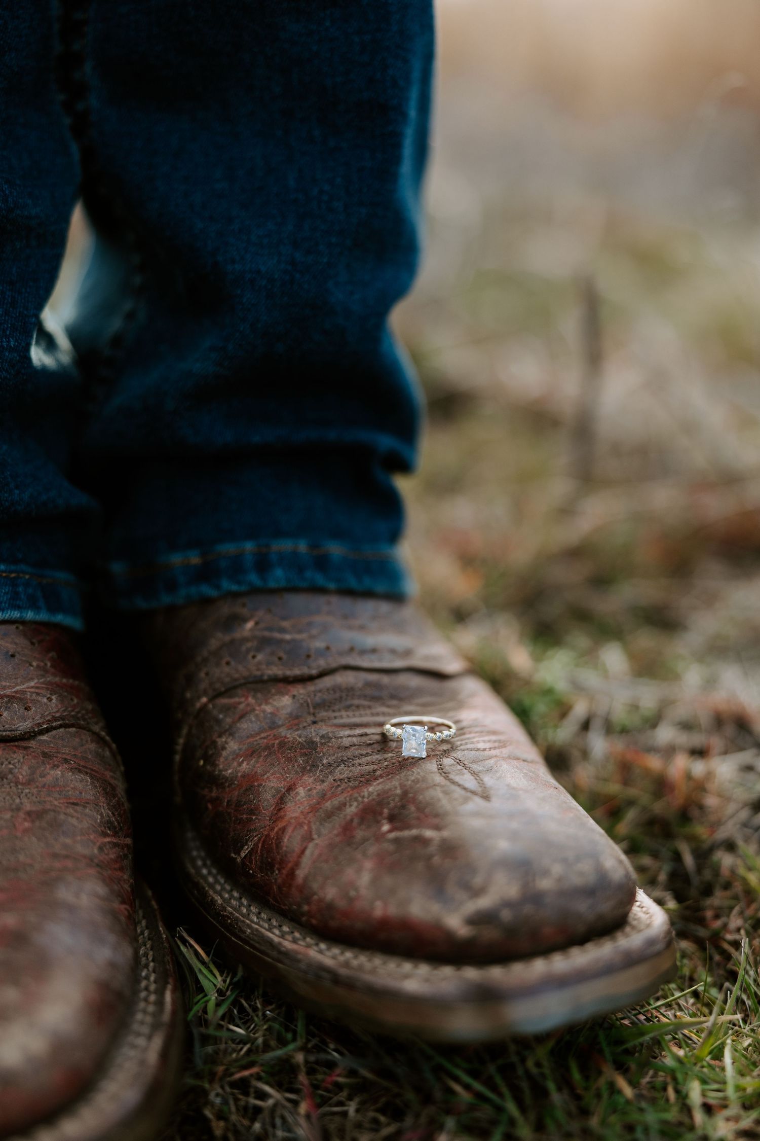 A diamond ring rests on muddy cowboy boots in grass.