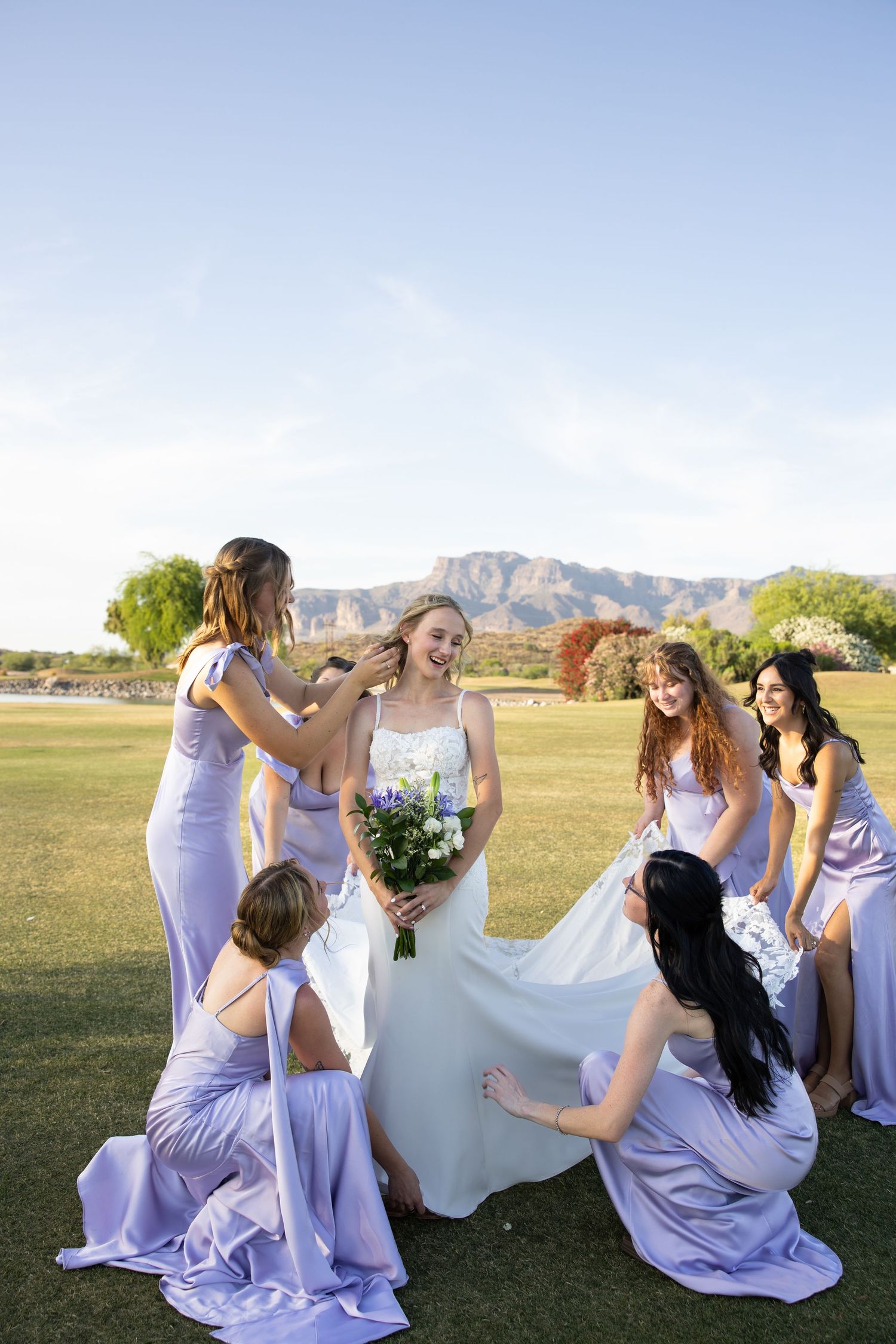 Bride in white gown holds bouquet surrounded by bridesmaids in lavender dresses on green lawn with mountain backdrop.