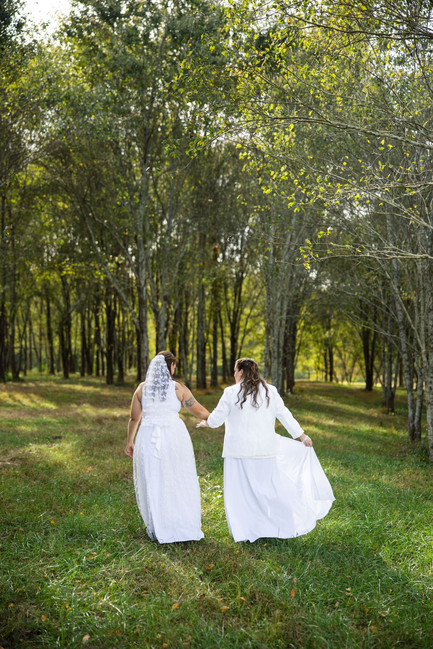 Two figures in flowing white dresses walk together through a sunlit forest path surrounded by green grass and trees.