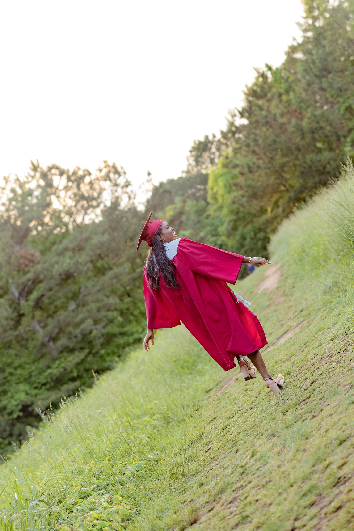 A graduate in red cap and gown poses playfully on a grassy hillside.
