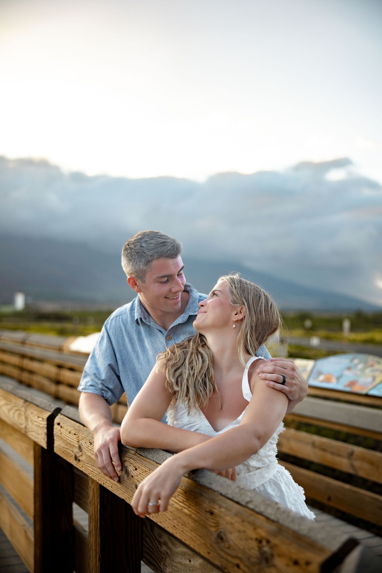 A romantic couple shares intimate moments on wooden benches with mountains in the background at sunset.