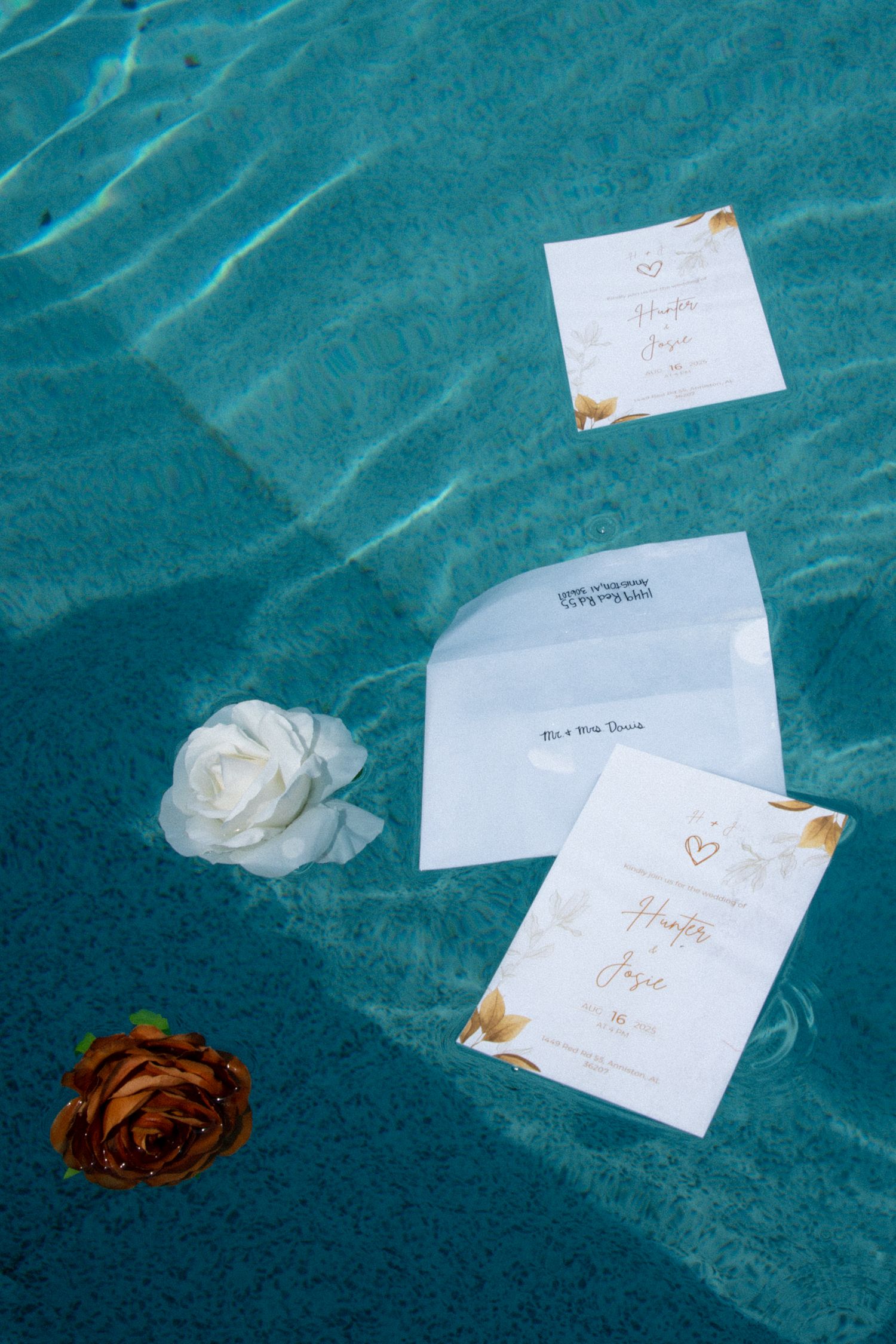 Wedding invitations and roses floating in a turquoise pool, featuring gold accents and elegant calligraphy.