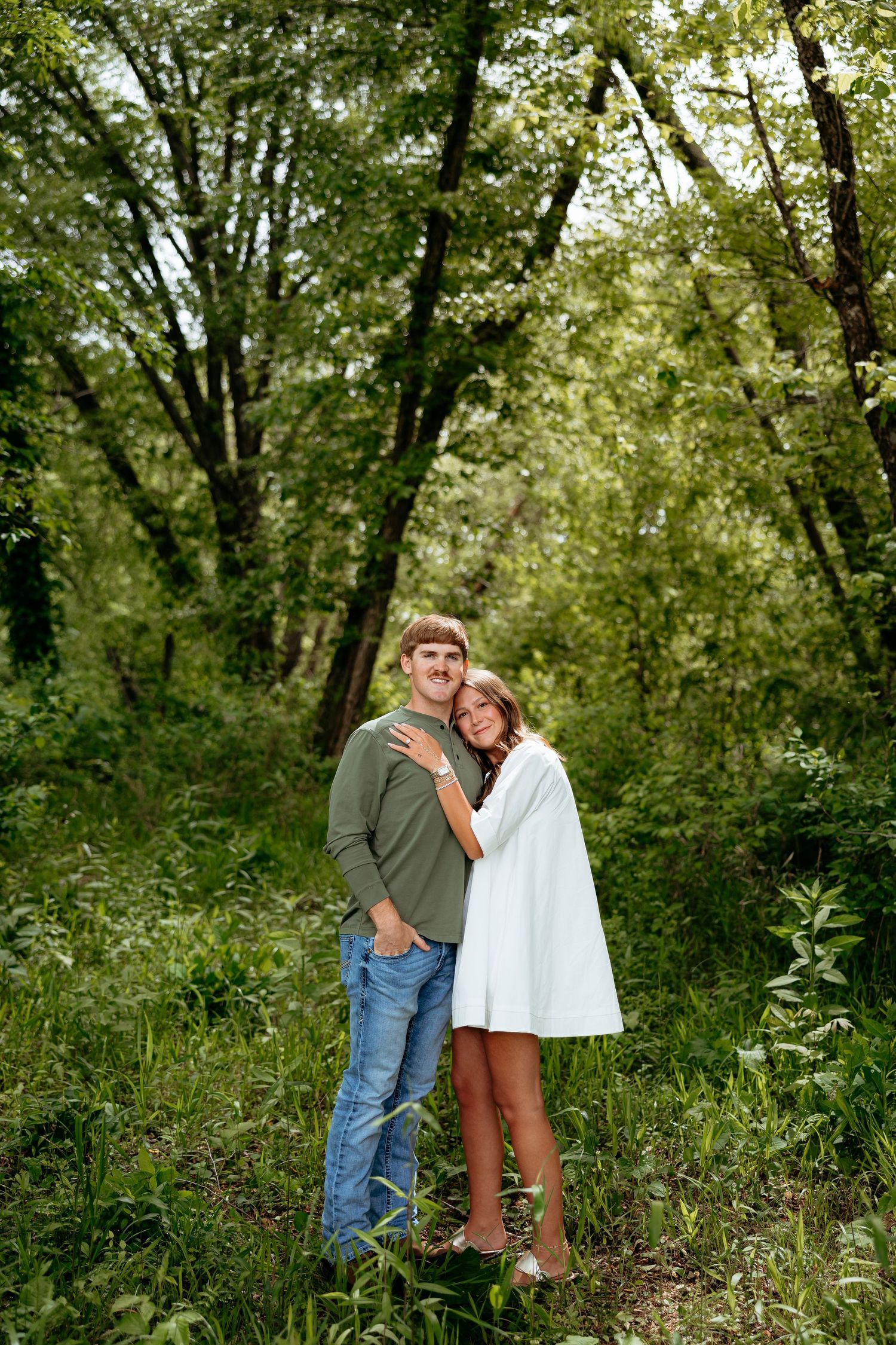 A couple embraces in a lush green forest, woman in white dress leans on man in green shirt and jeans.