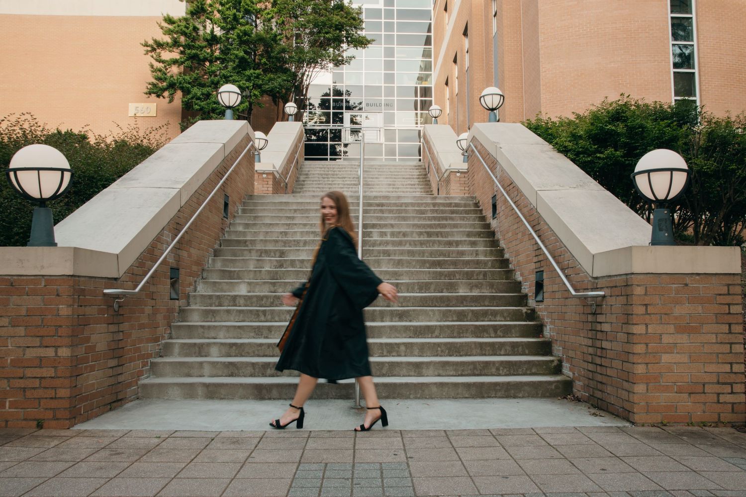Someone in a flowing dark dress and heels twirls playfully on a brick staircase with decorative lampposts.