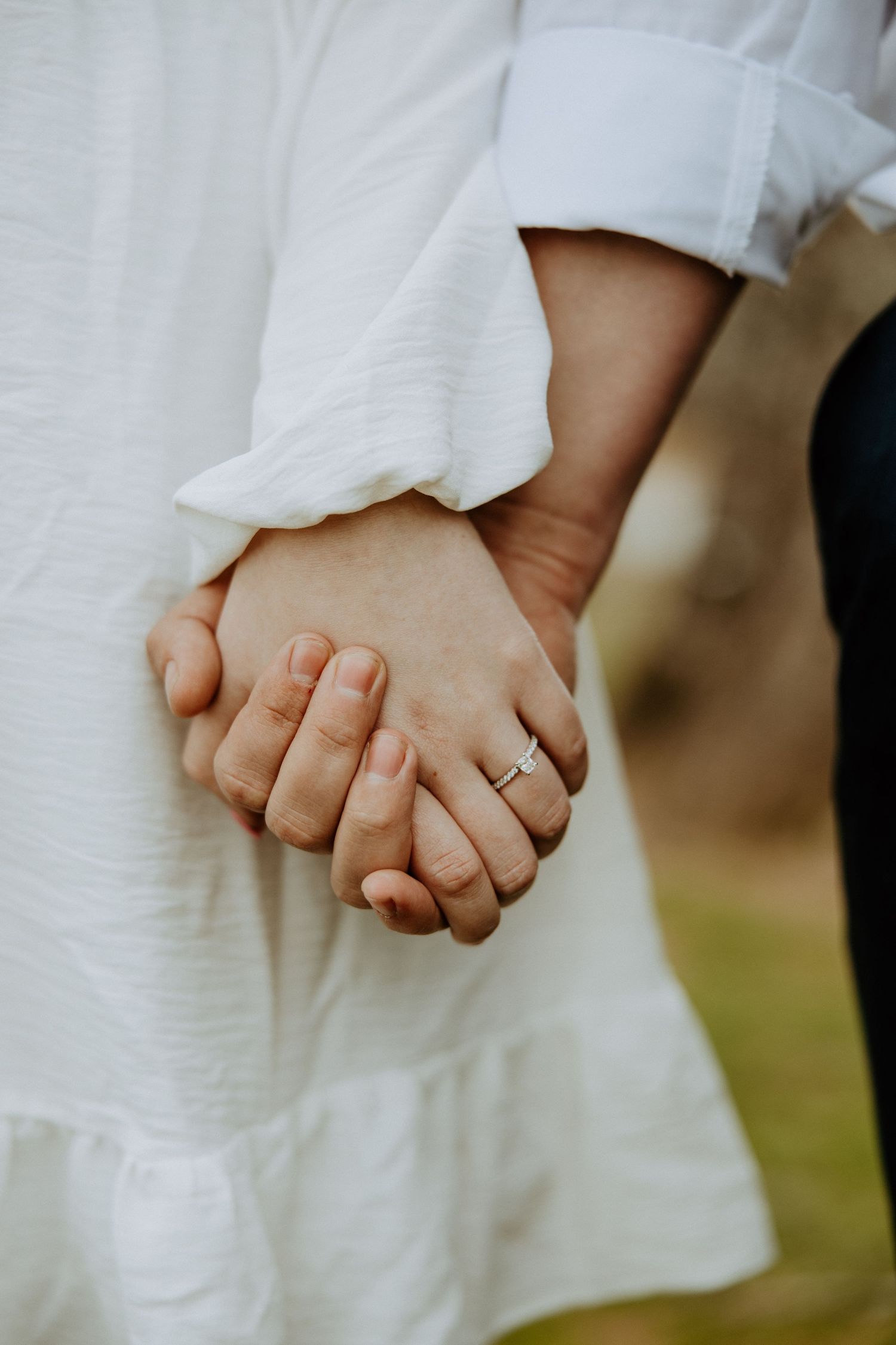 Close up of two hands clasped together with an engagement ring visible against a white dress background.