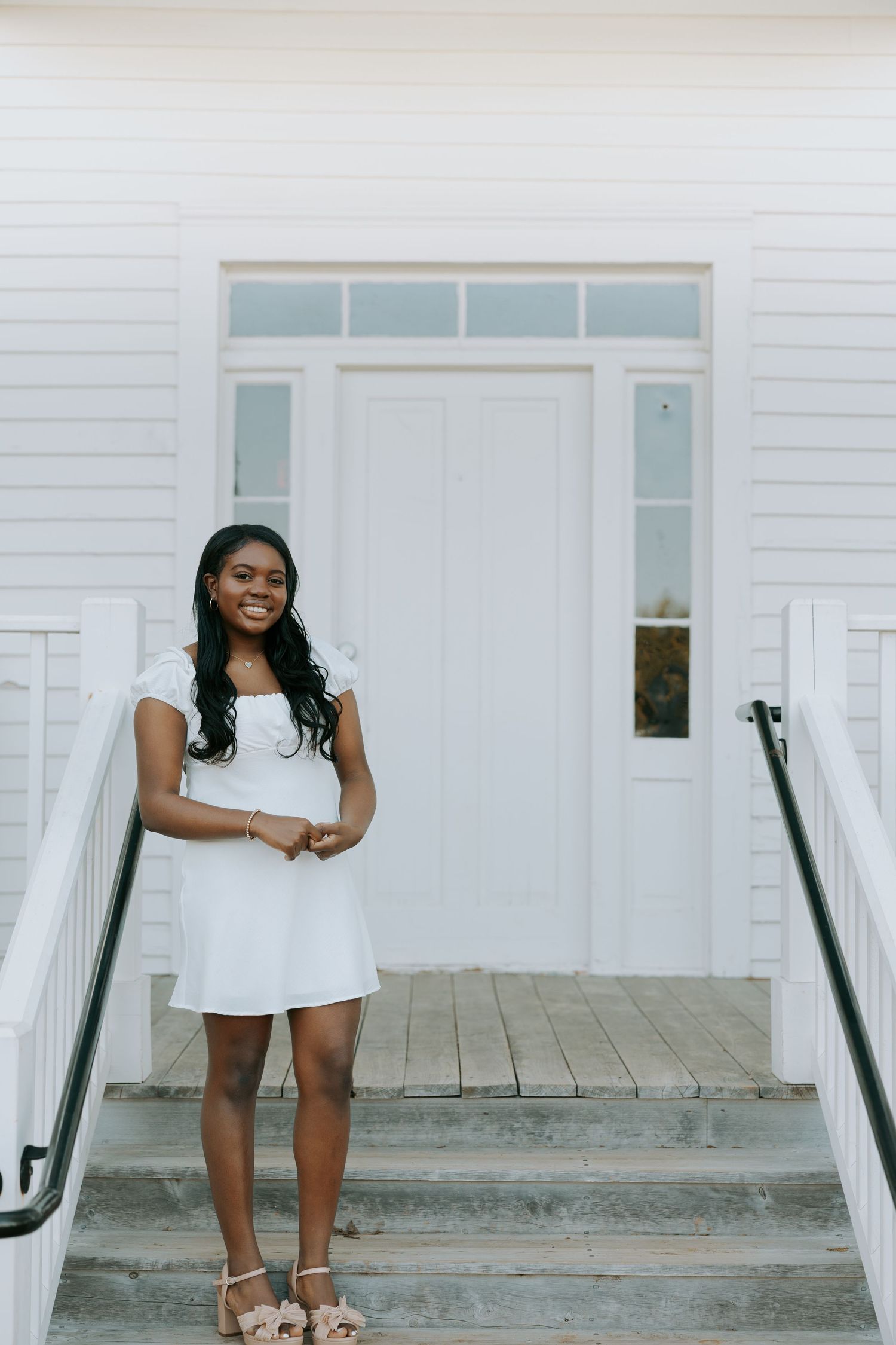 A person in a white dress poses on steps leading to a white doorway with transom windows.