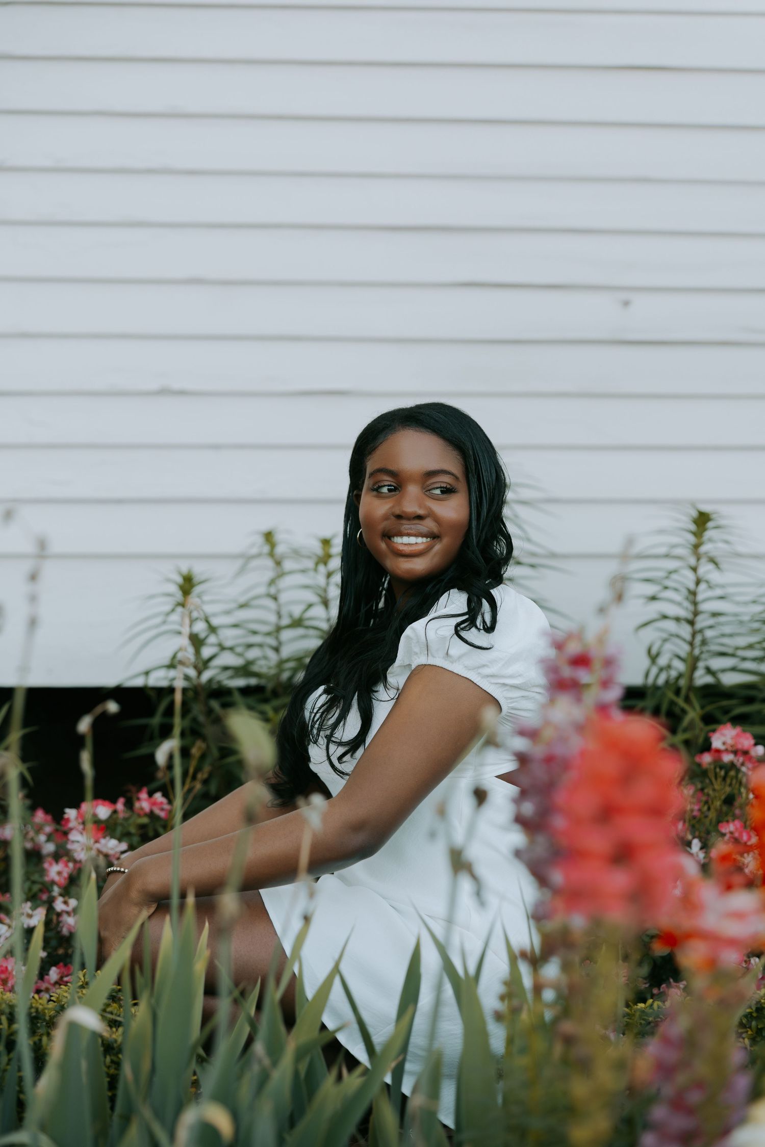 Person smiling among colorful garden flowers against white siding.