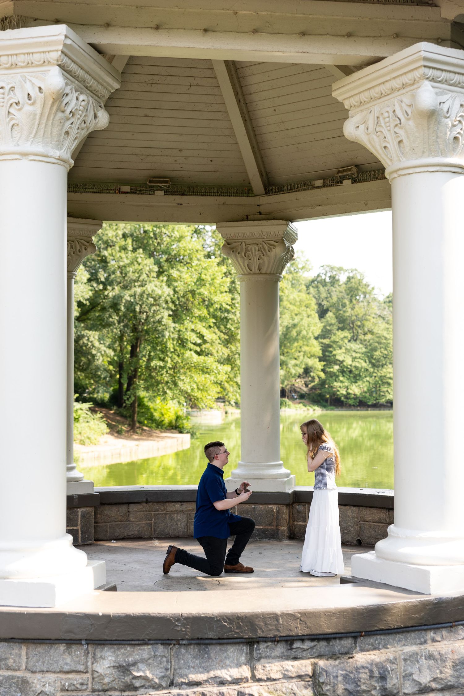 Man proposes on one knee under elegant white columns of a gazebo overlooking a serene pond surrounded by lush greenery.