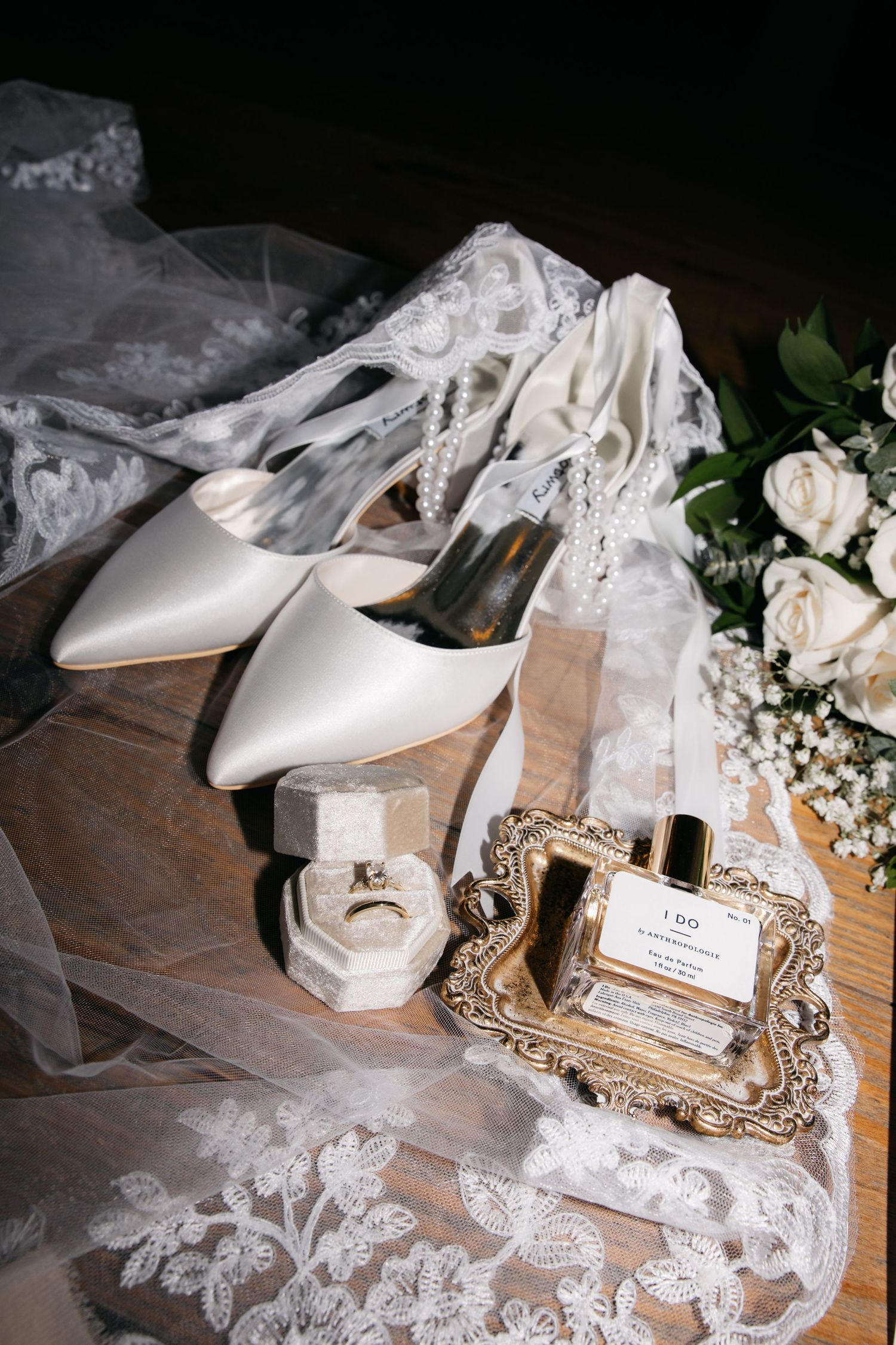 White wedding shoes and delicate lace veil arranged with floral accents on rustic wooden surface.