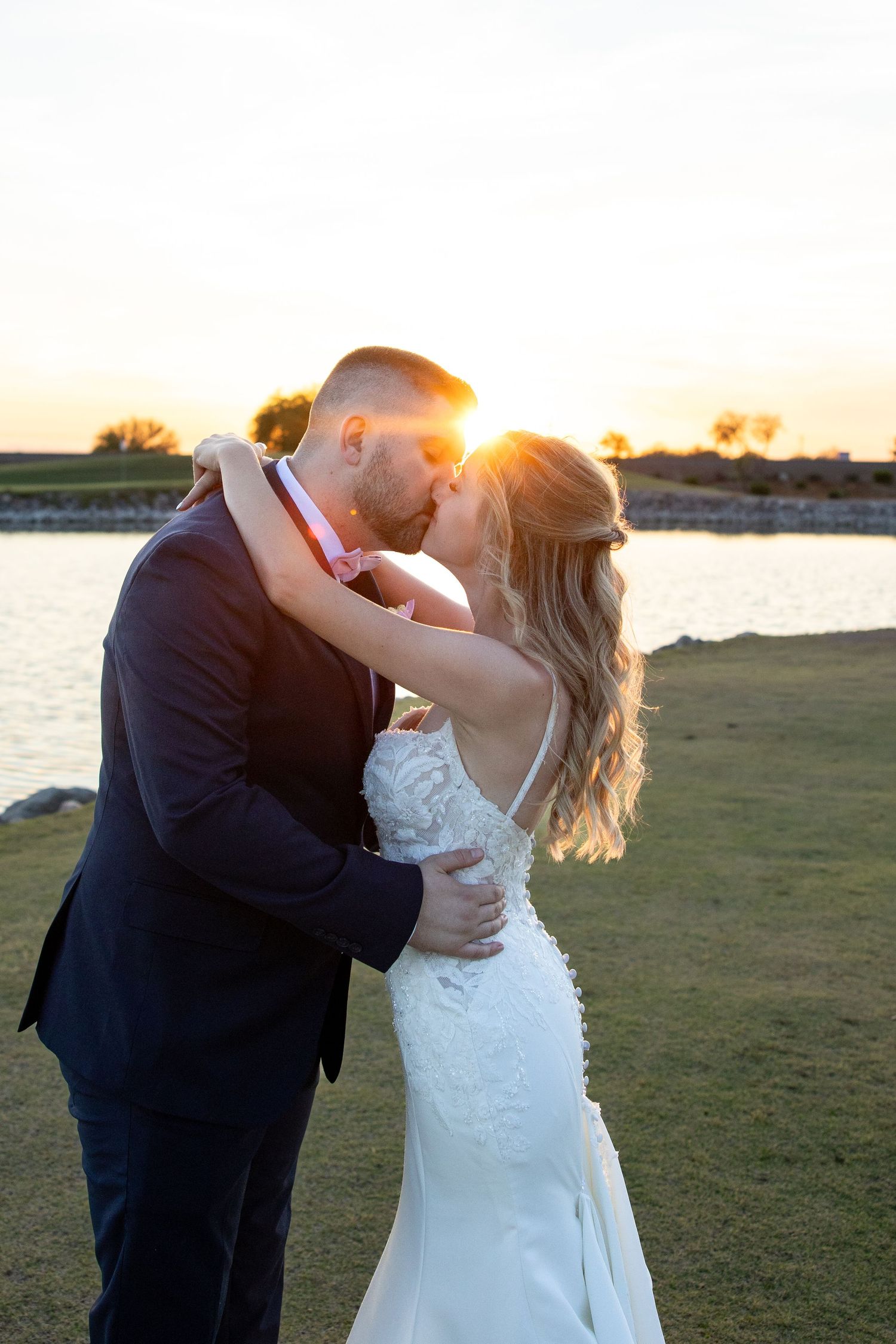 Bride and groom sharing a romantic kiss at sunset with a lake and golden sky behind them.