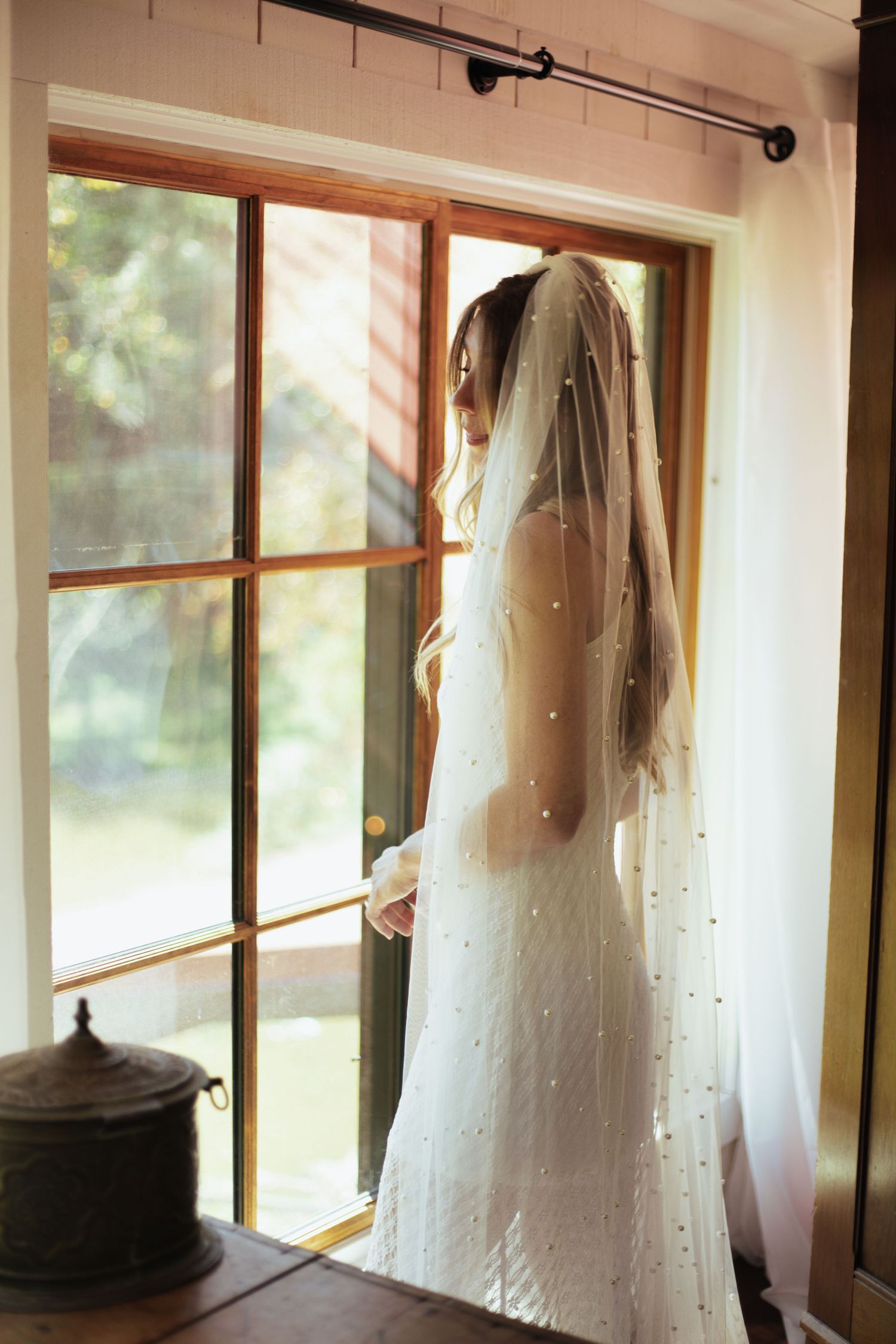 Sunlit silhouette of a bride in her wedding dress standing by a window.