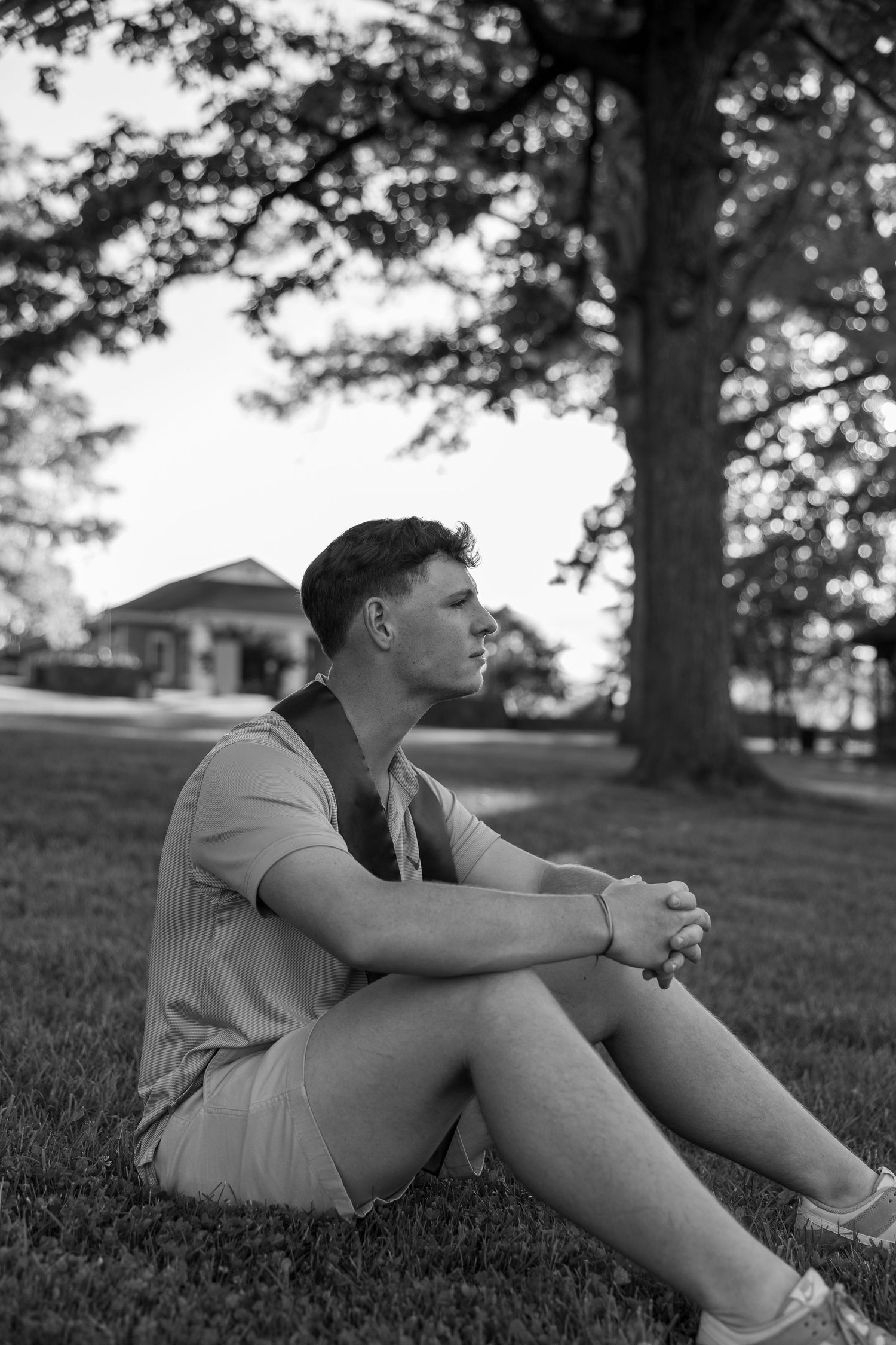 Black and white portrait of person sitting on grass near a large tree.