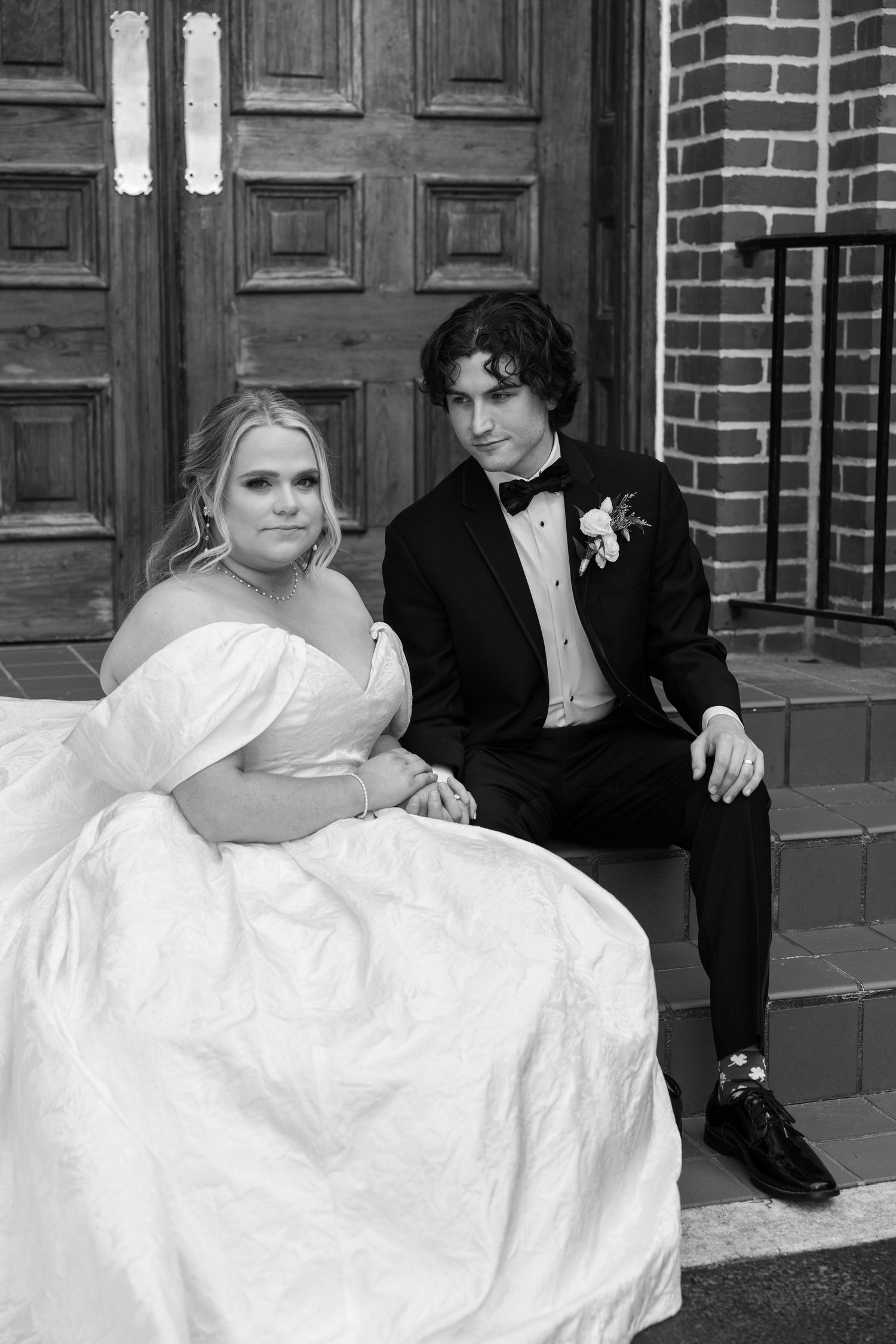 Black and white photo of a bride in a white gown and groom in a tuxedo sitting on church steps.