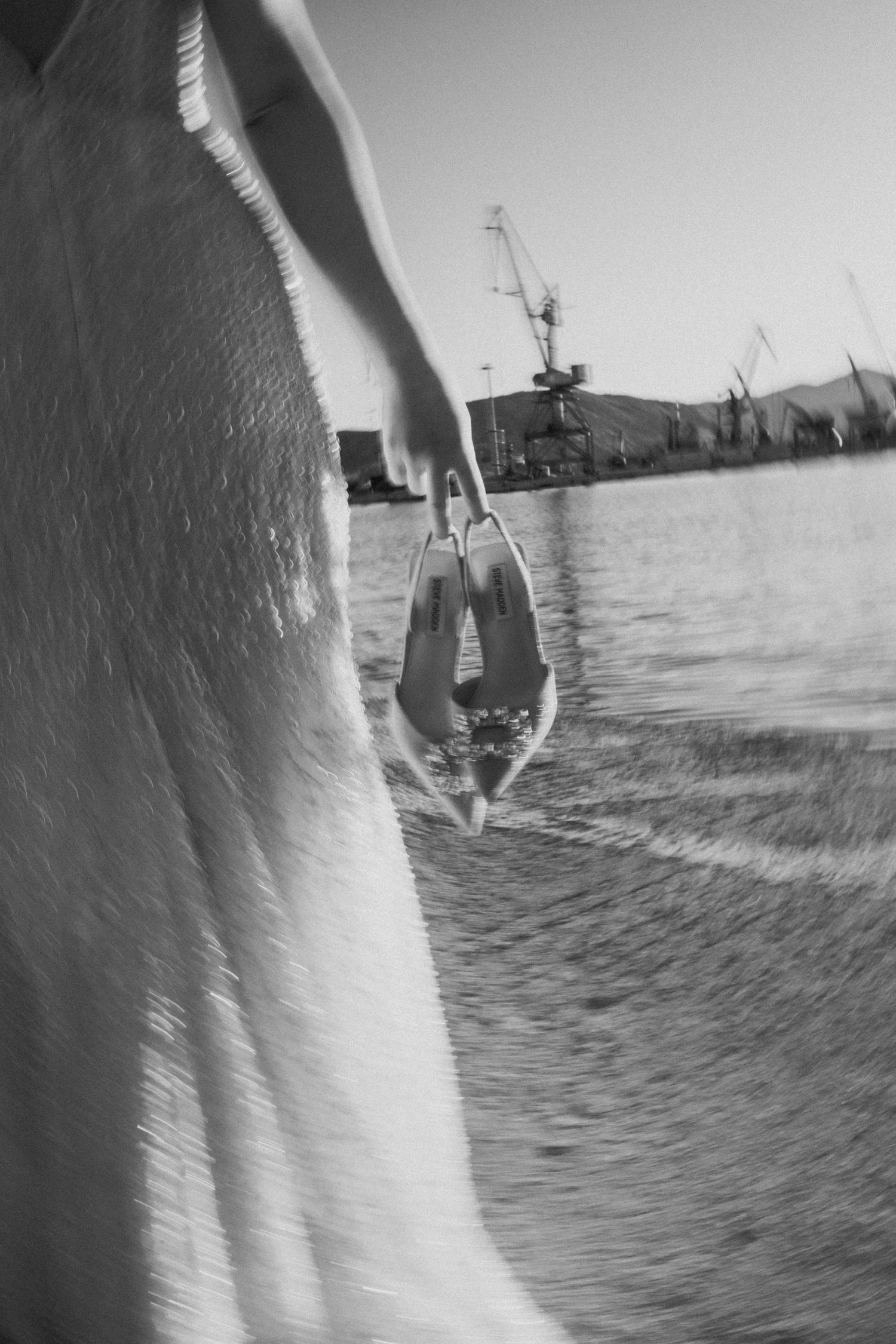 fine art φωτογραφια ασπρομαυρη με την νυφη να κραταει τις νυφικες γοβες στα χερια στο λιμανι του βολου