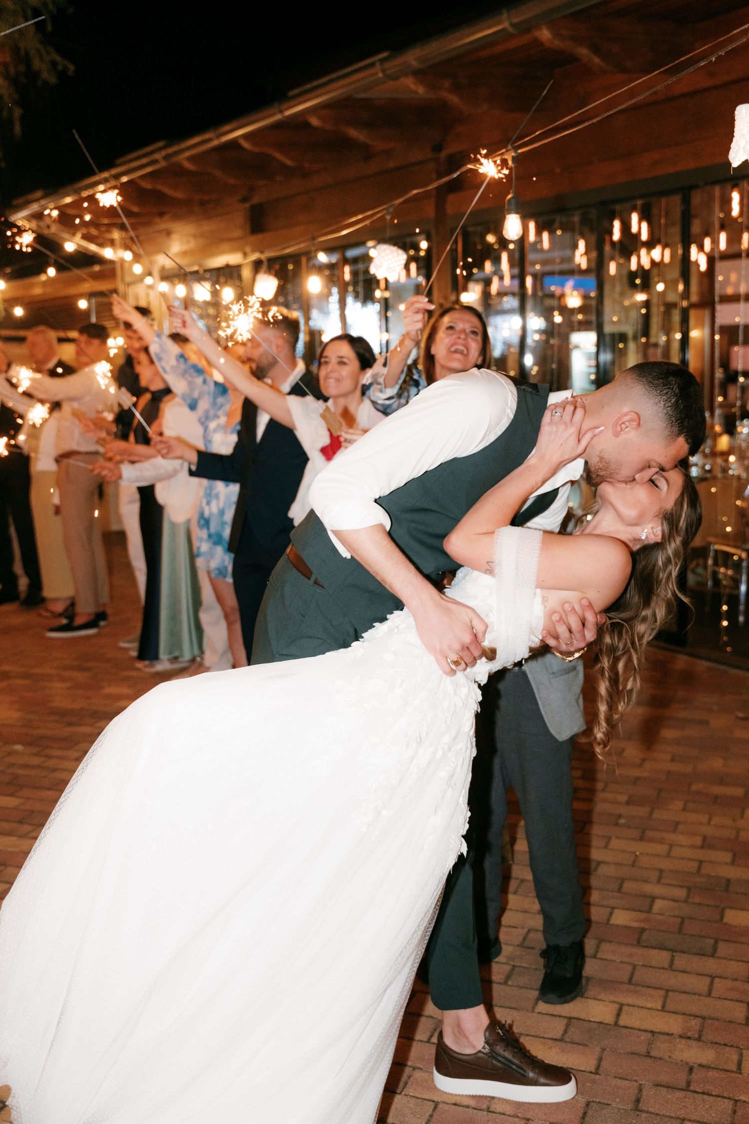 Groom dips bride for a dramatic kiss while wedding guests wave sparklers in celebration at outdoor reception. Mallorca wedding photographer