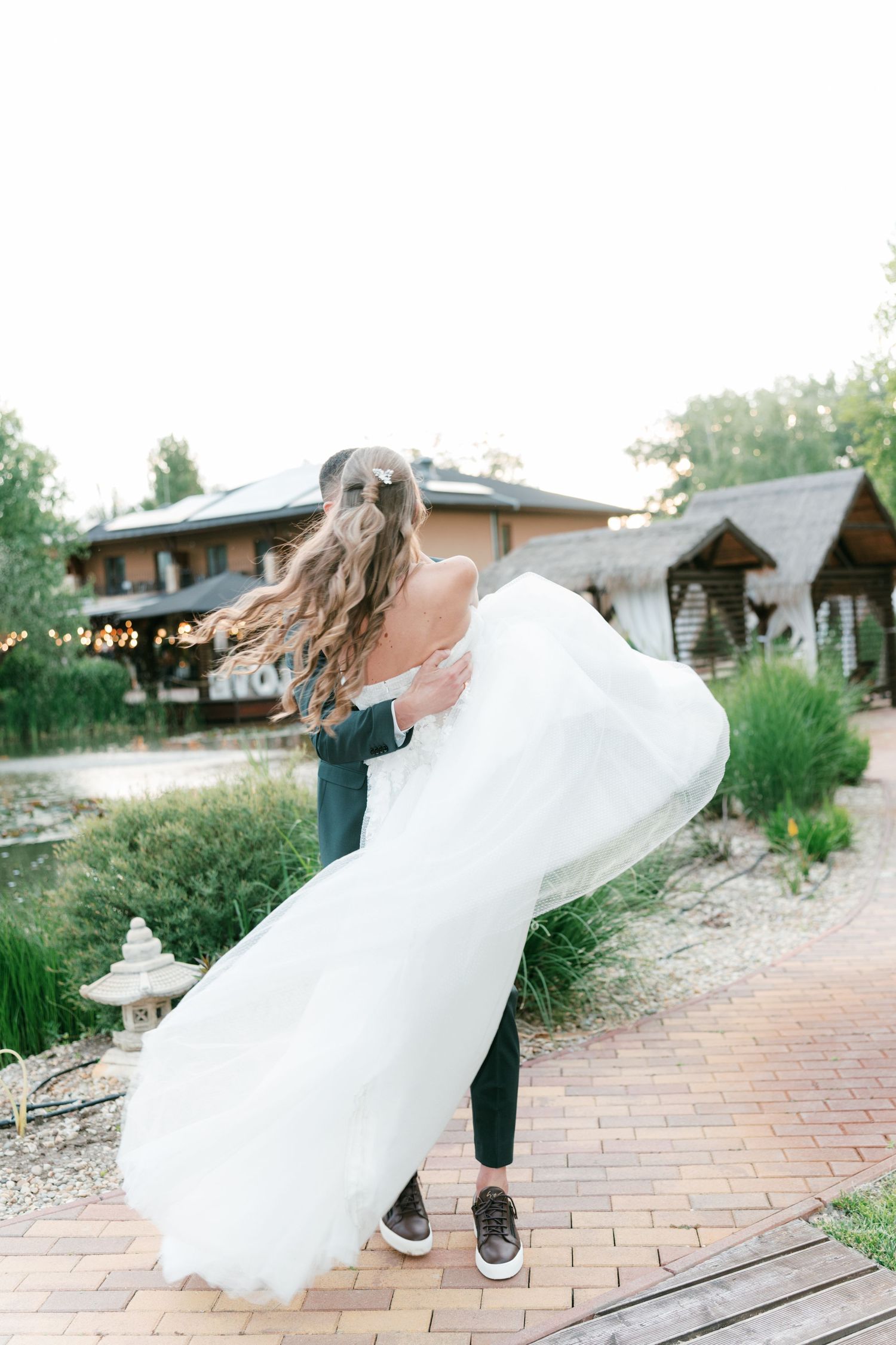 Groom lifts and spins bride in white gown outdoors at rustic lakeside wedding venue. Mallorca wedding photographer and filmmaker.