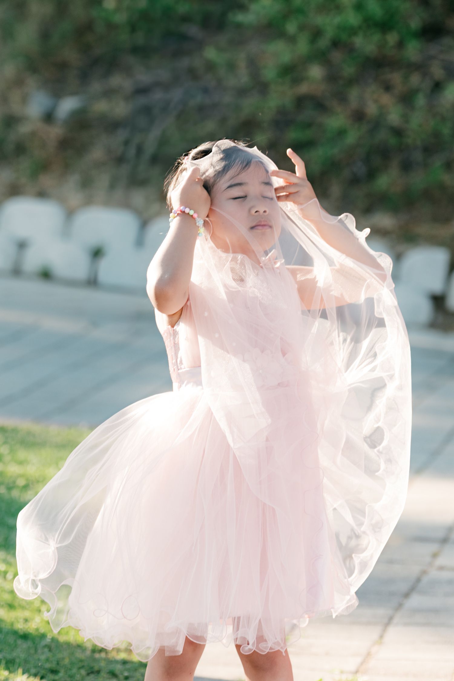 Young girl in pink tulle dress adjusts veil outdoors at sunny wedding venue. Italy wedding photographer and filmmaker