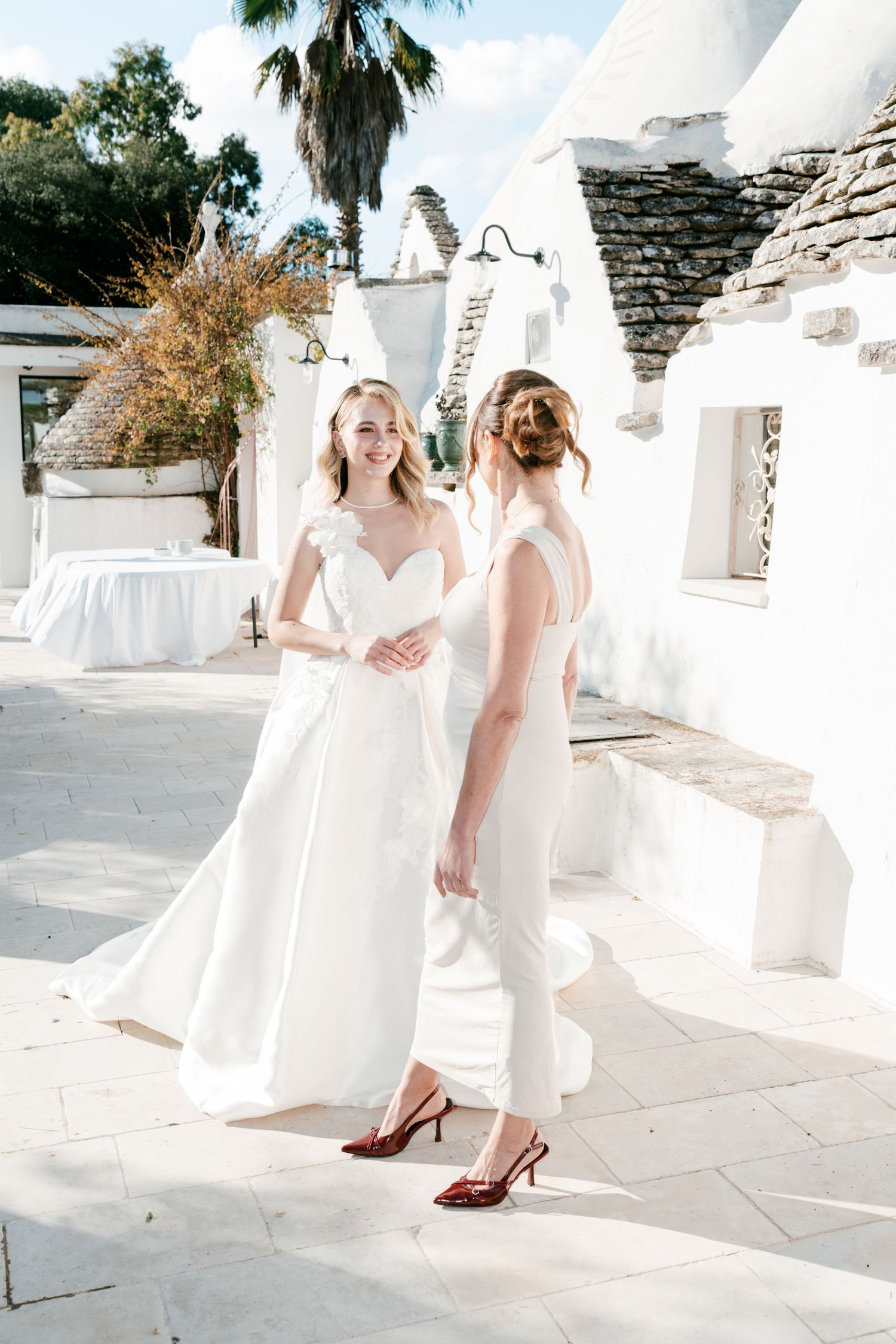 Smiling bride in white gown with bridesmaid in white jumpsuit walking past traditional trulli buildings. Puglia wedding photographer, film