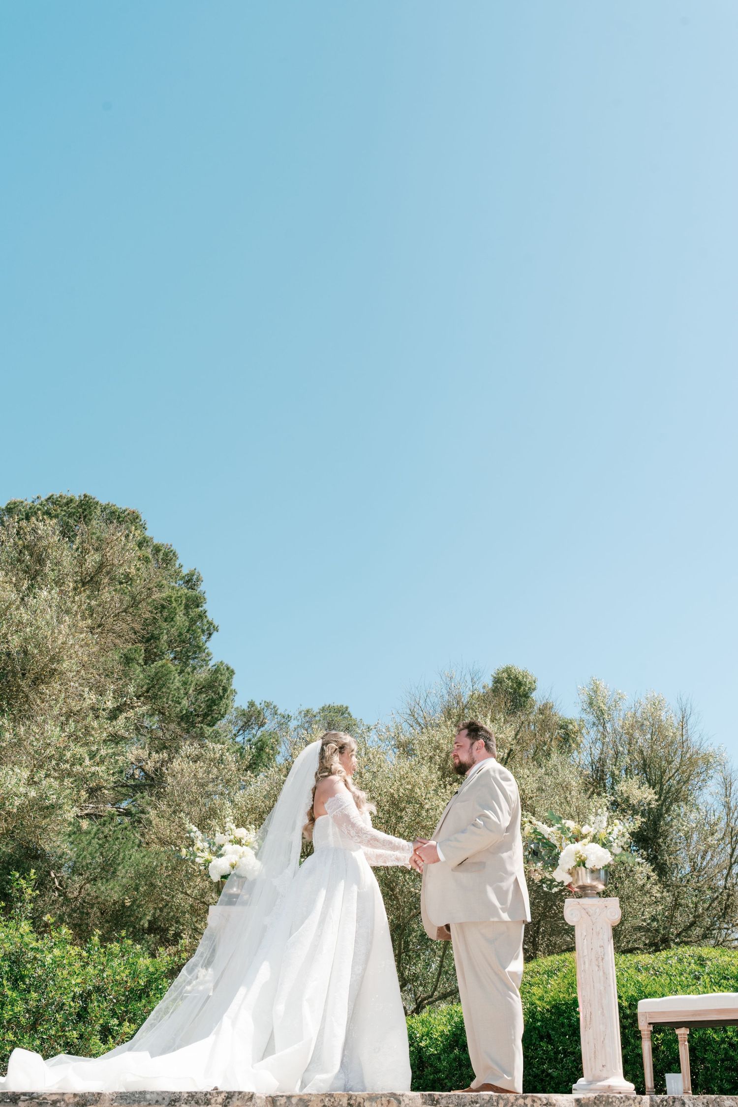 Bride and groom holding hands at outdoor wedding ceremony under blue sky surrounded by lush green trees.