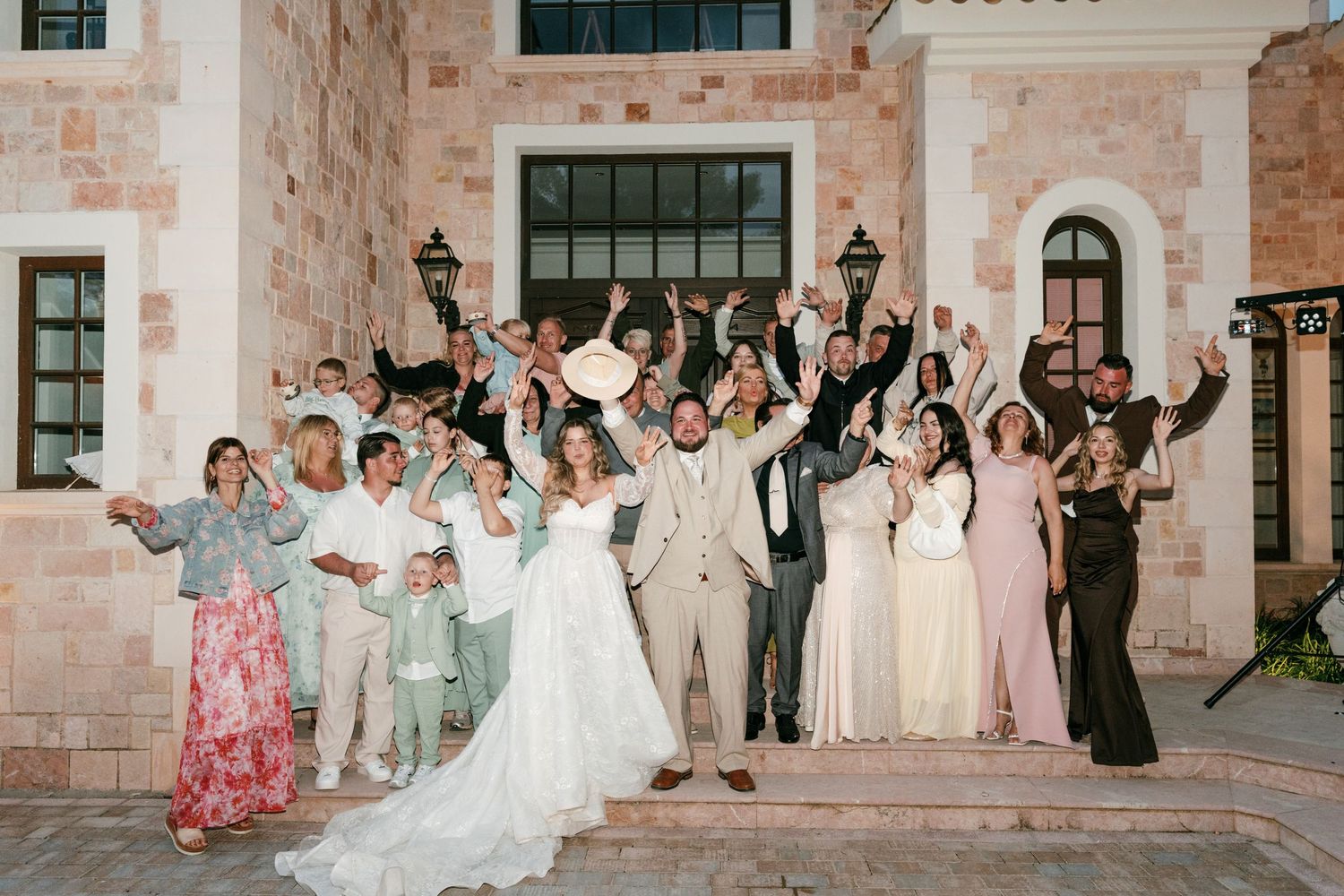Wedding party celebrating outdoors at night, raising hands joyfully in front of a stone building, bride in white gown centered.