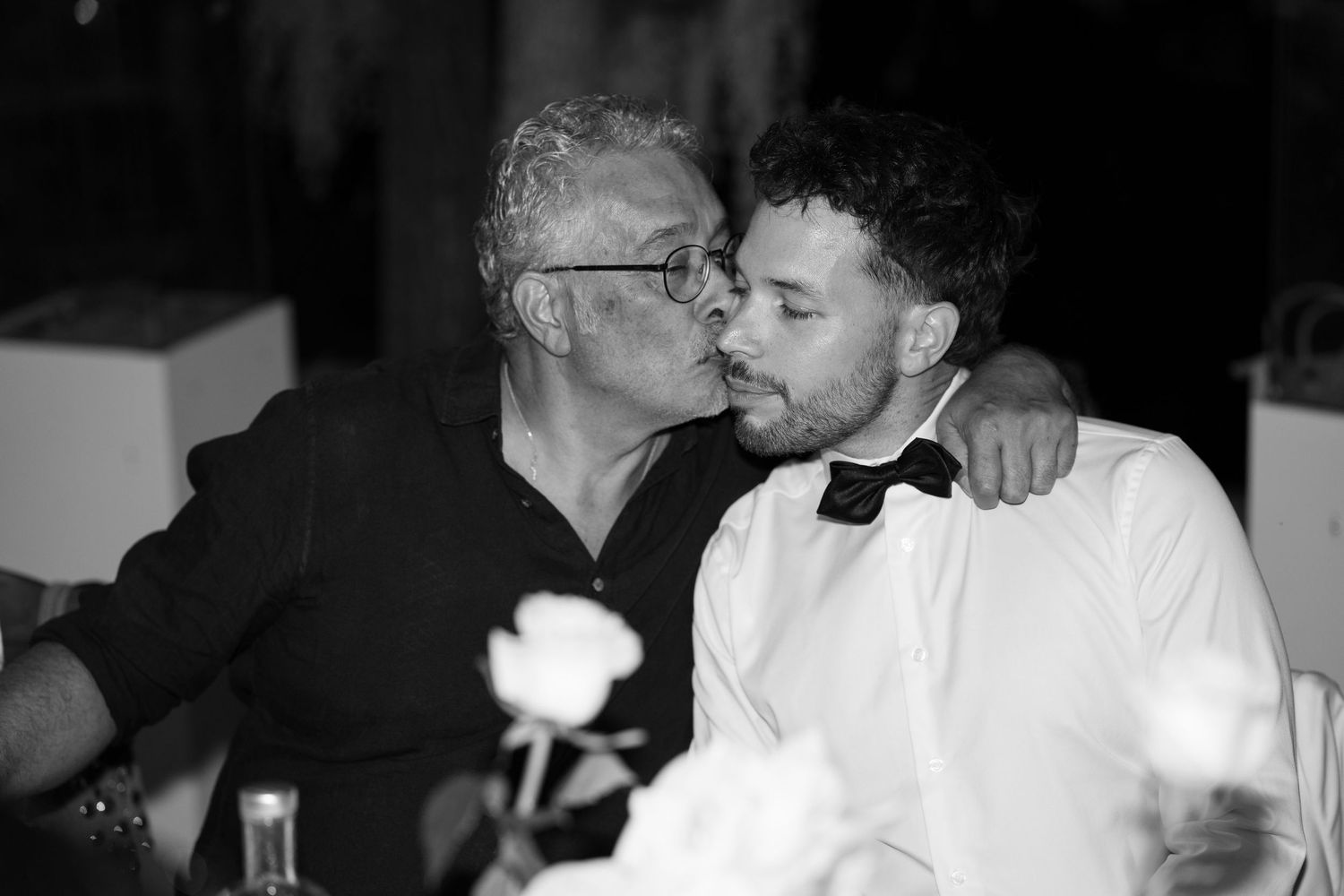 Black and white photo of two men sharing a kiss at a formal event, one wearing a bow tie.
