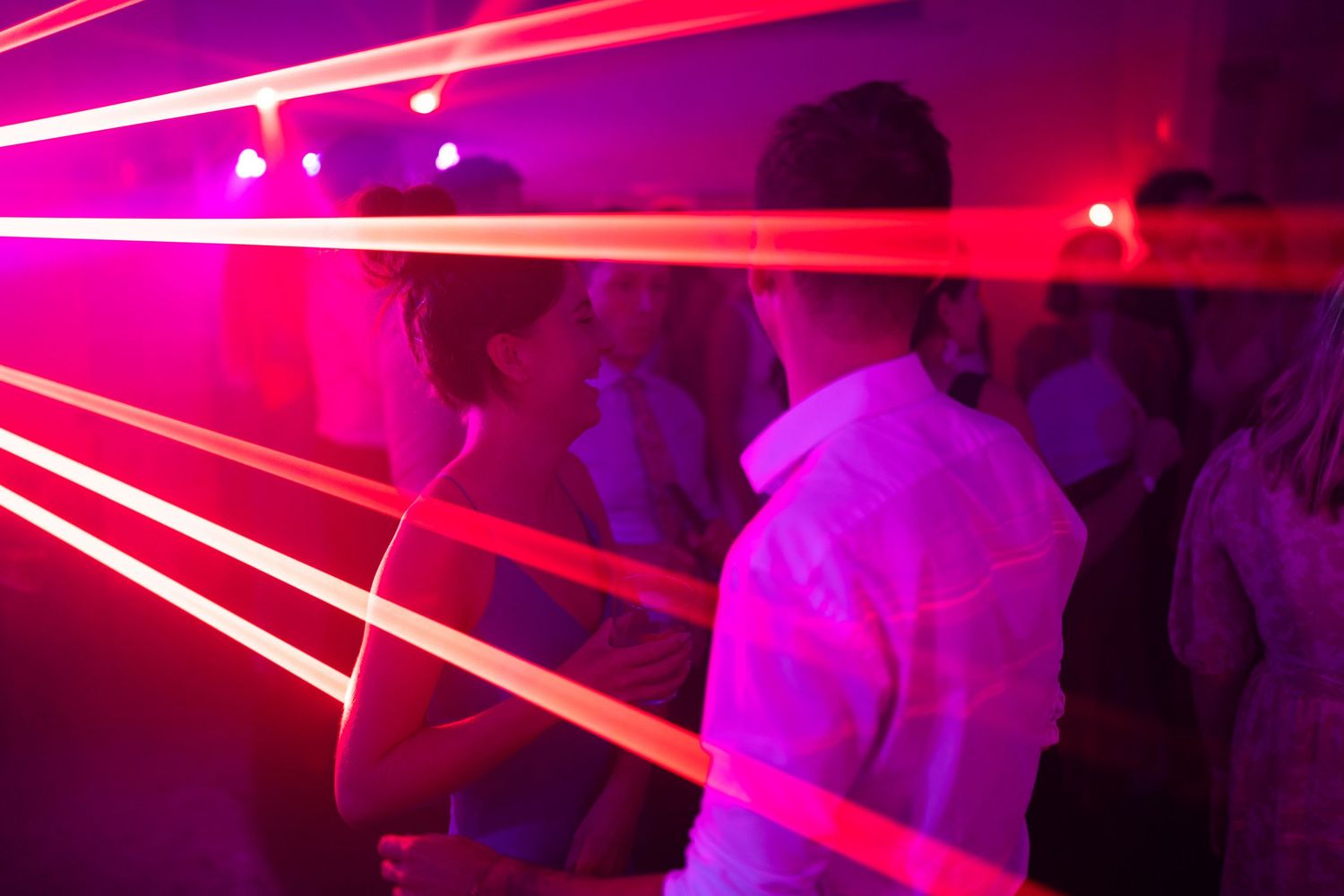 Guests dance in a nightclub as vivid pink and red laser beams stream across the room, lighting a man in a white shirt and a woman.