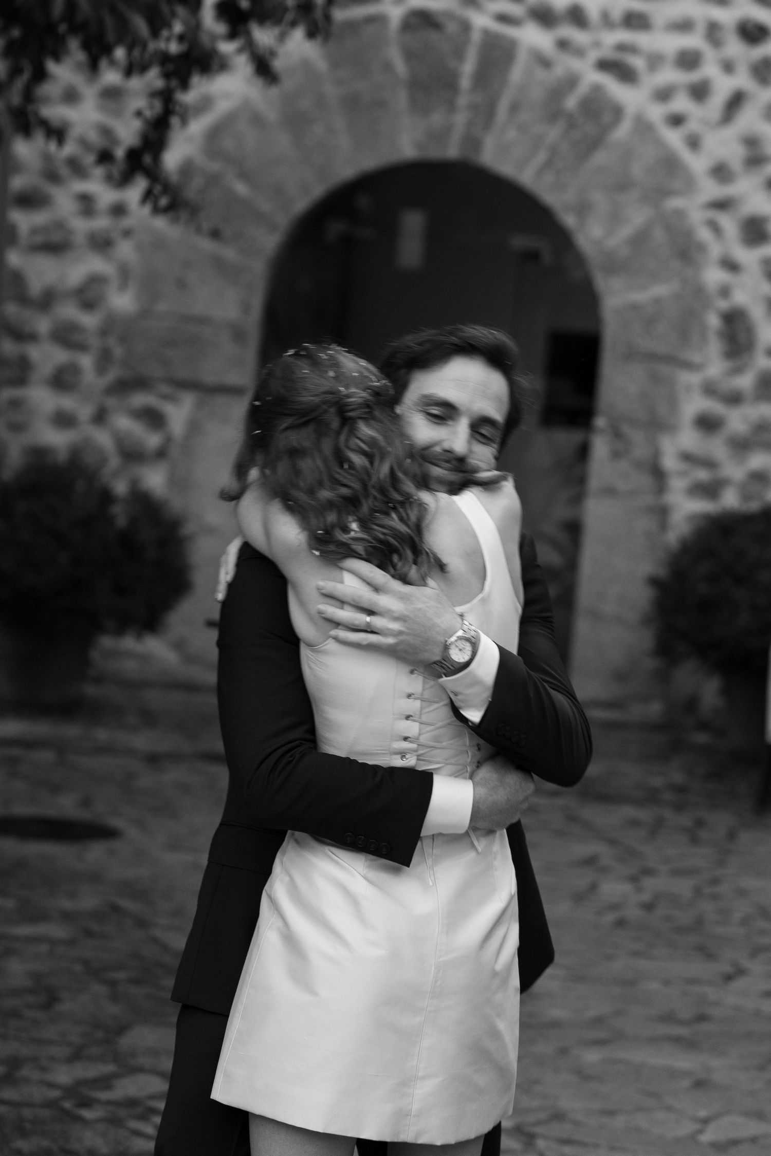 Black and white photo of a smiling man in a dark suit embracing a woman in a short white dress under a stone archway.