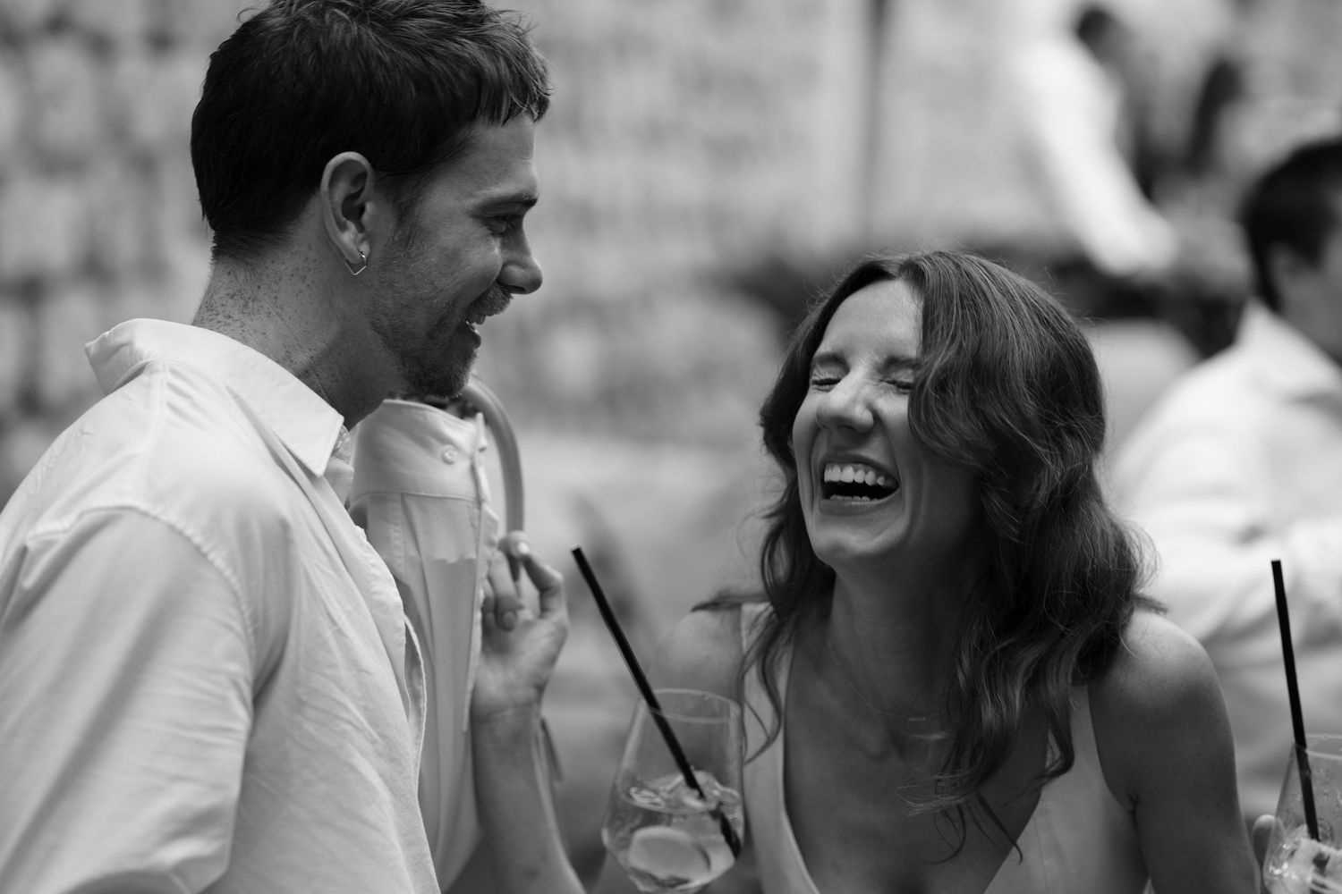 Black and white photo of a smiling man and a laughing woman holding drinks with straws at a lively outdoor gathering.