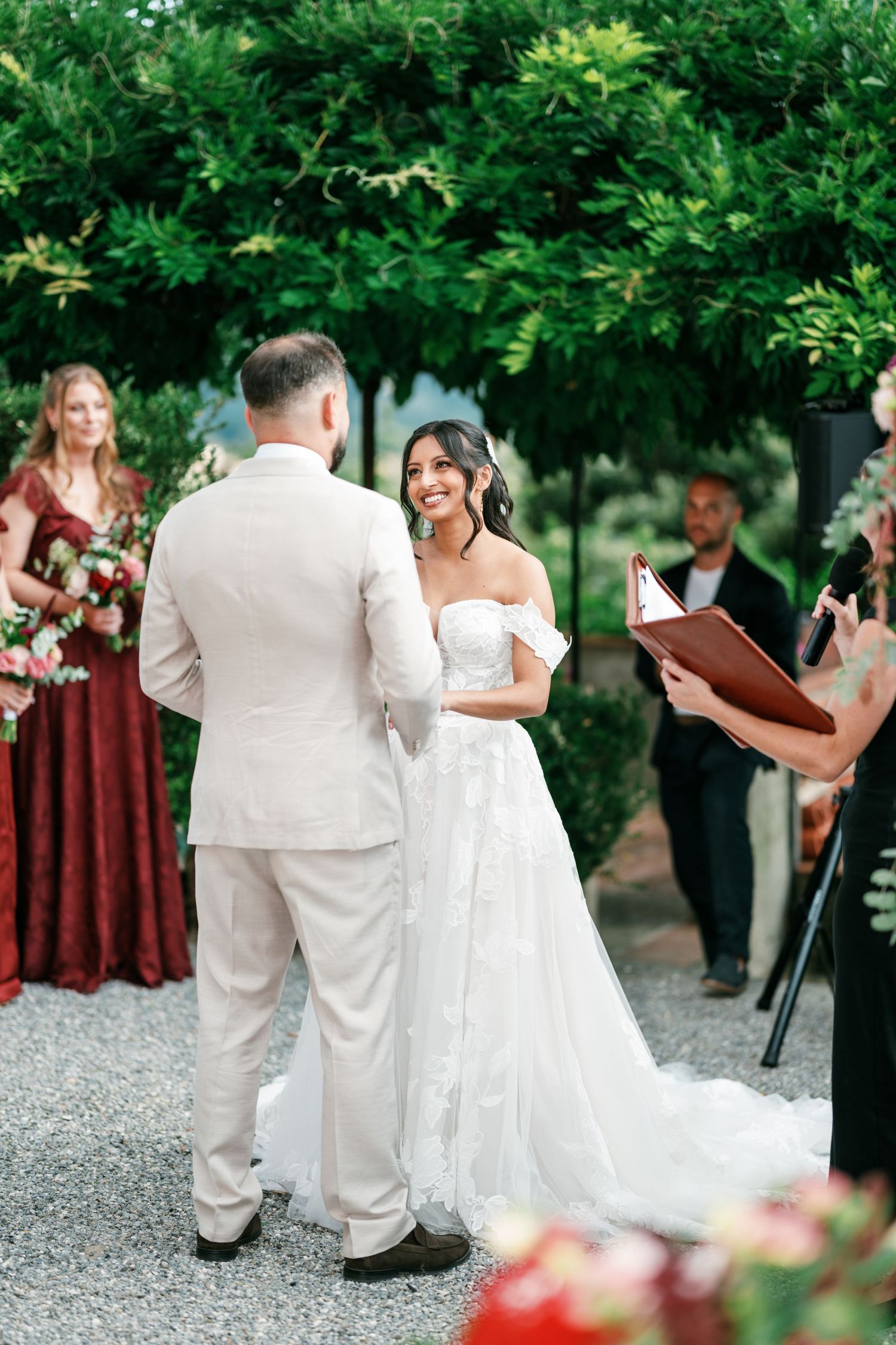 Smiling bride in a white off-shoulder lace gown faces the groom in a beige suit during an outdoor garden wedding ceremony.