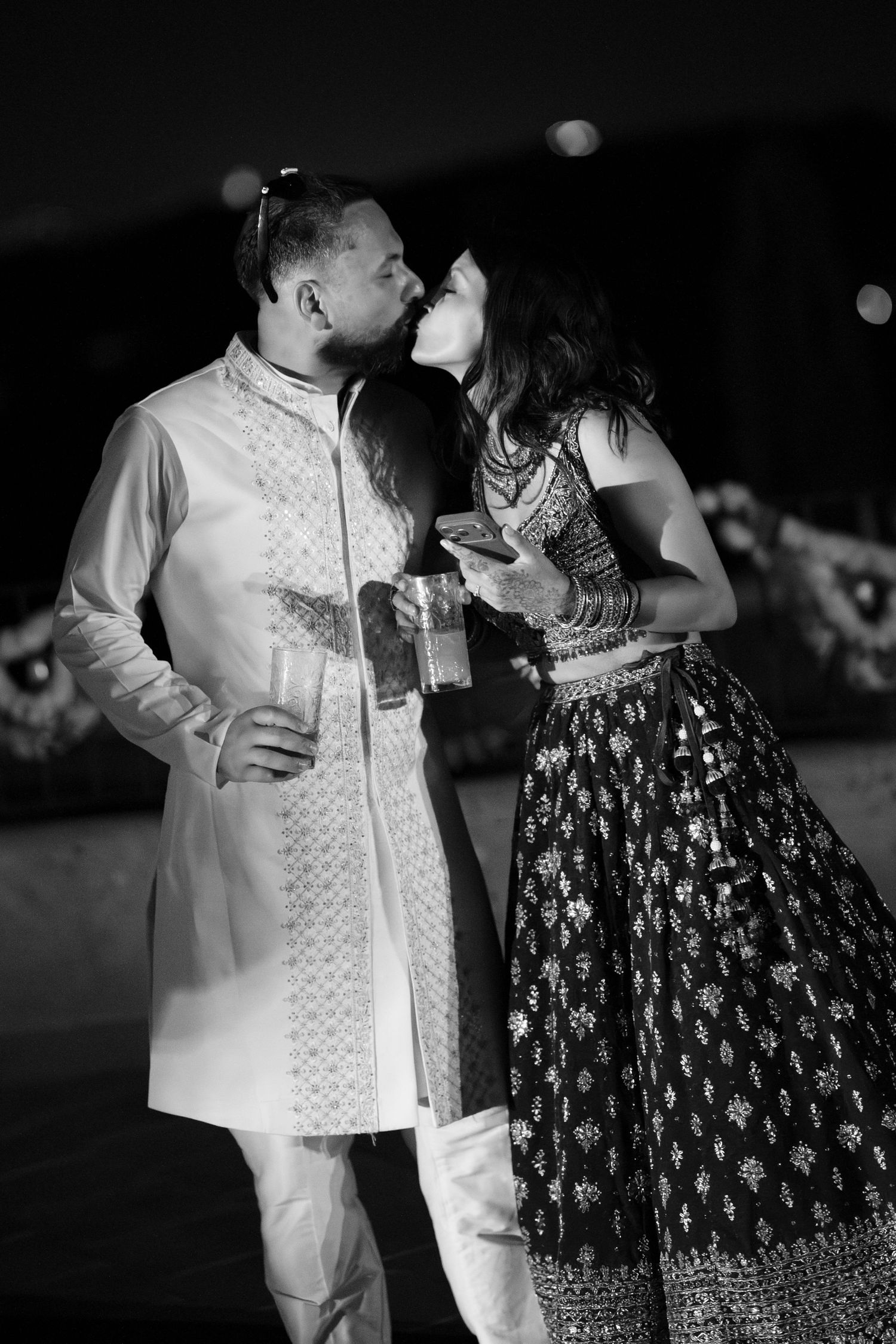 Black and white photo of a couple sharing a kiss at an Indian wedding celebration, he in a sherwani and she in a printed lehenga.