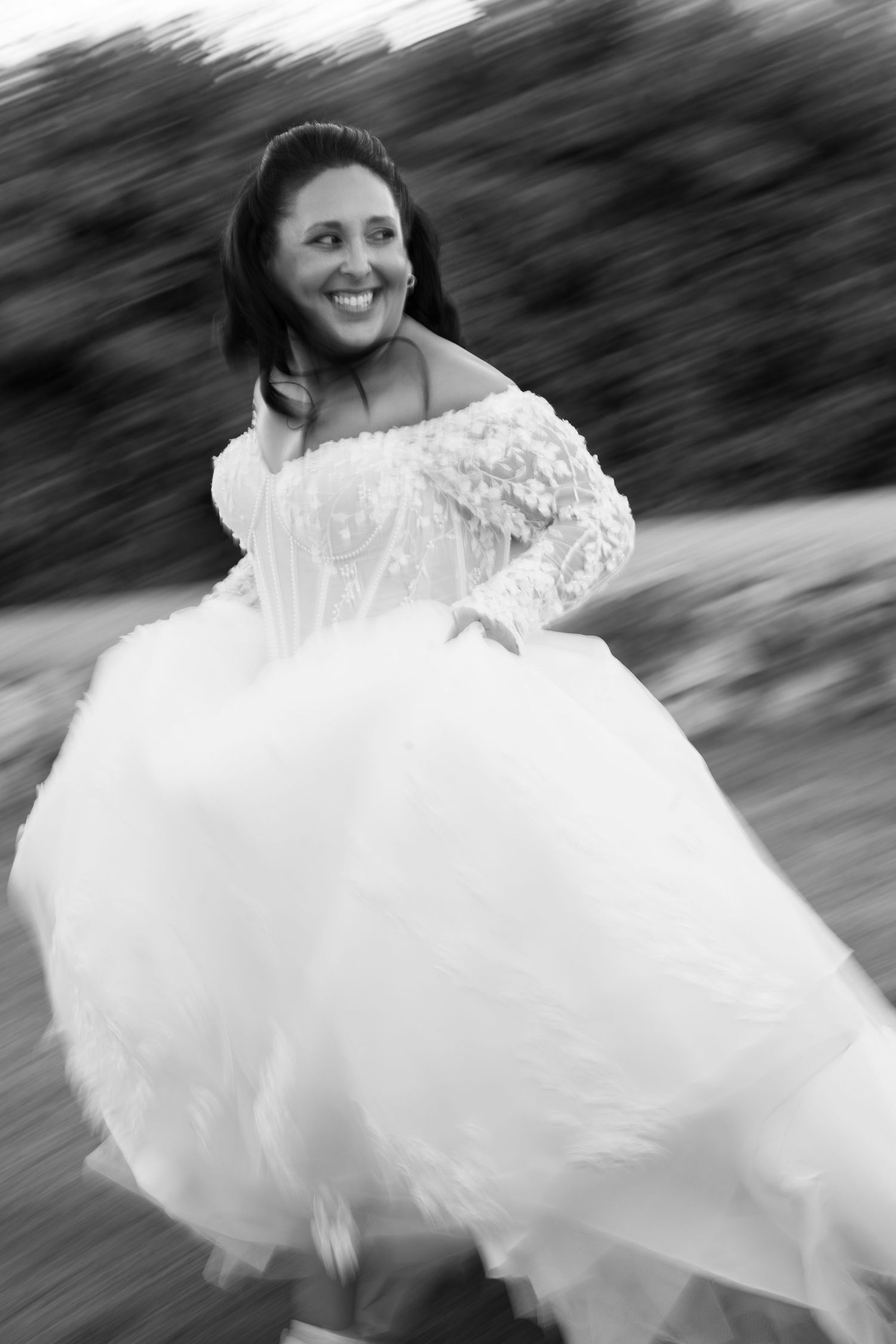 Black and white motion-blurred portrait of a smiling bride in a flowing tulle wedding gown with lace sleeves twirling outdoors.