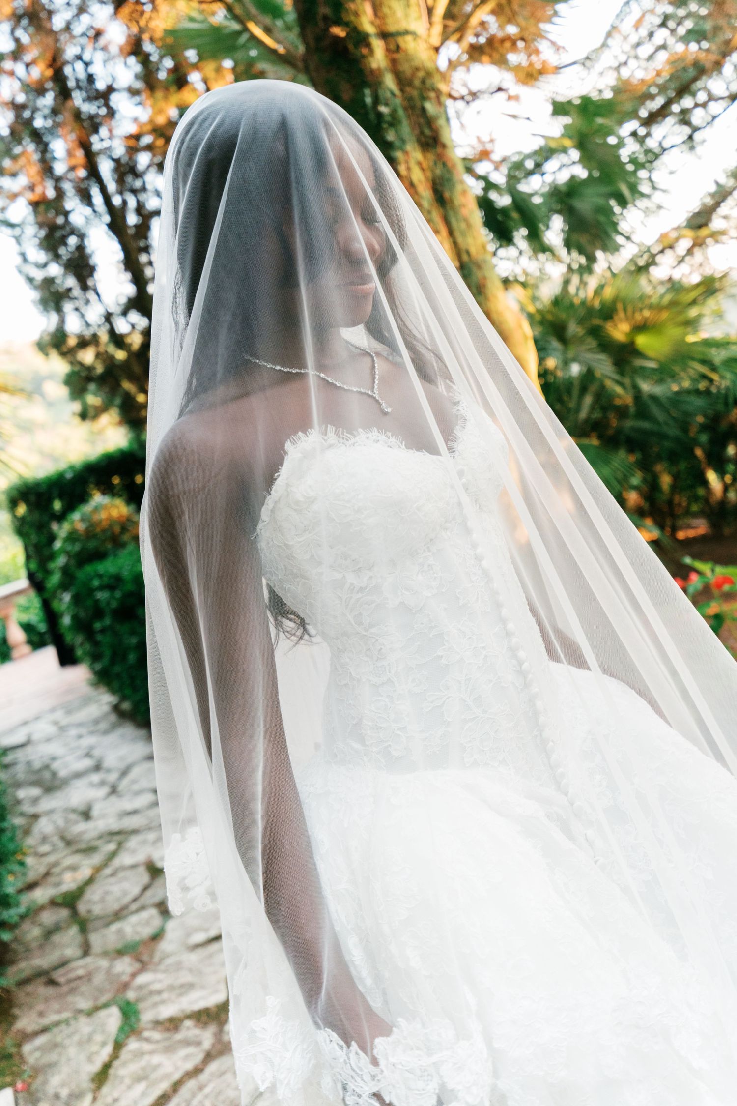 Bride in a strapless lace wedding gown covered by a sheer veil walks along a stone garden path beneath autumn trees.
