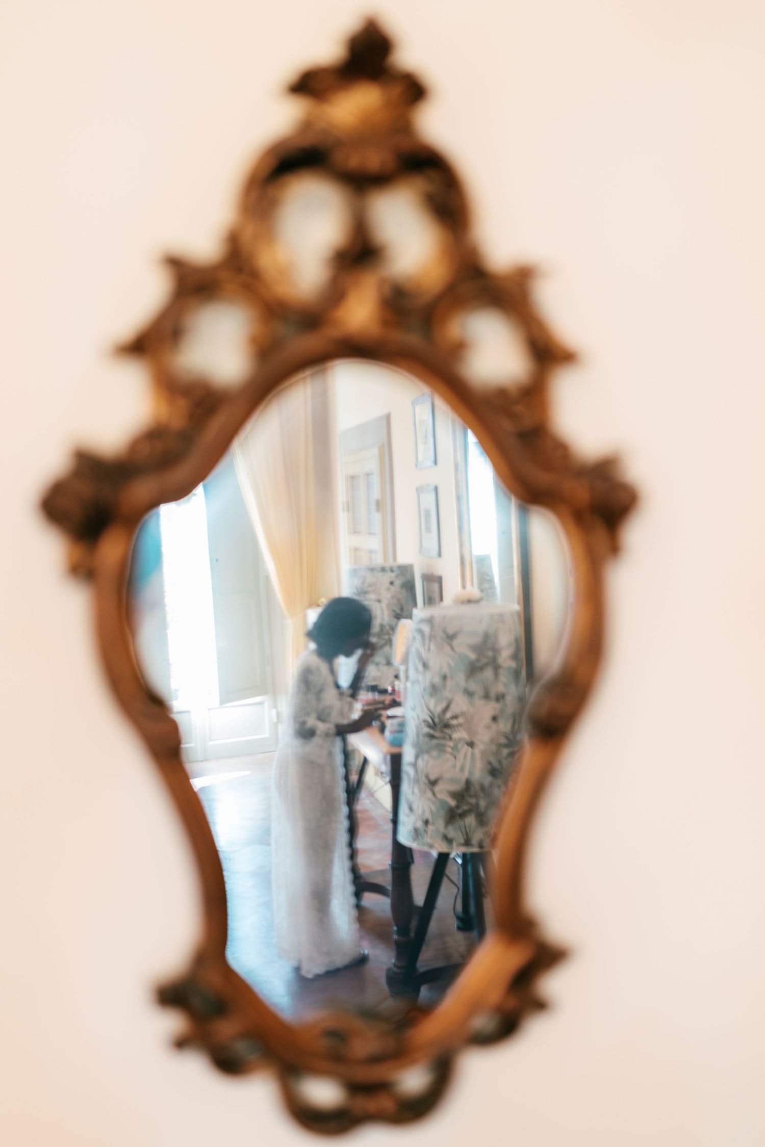 Ornate gilded antique mirror reflecting a bride in a white lace gown standing in a bright room with floral upholstered furniture.