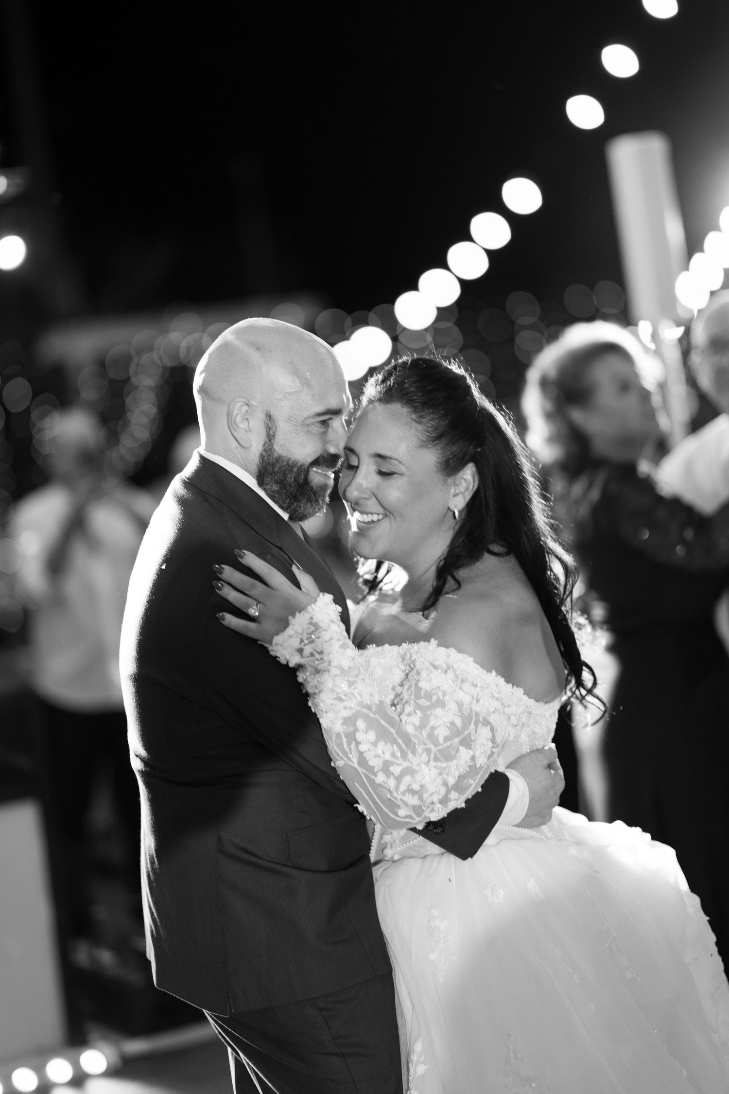 A bride and groom share a joyful embrace during their first dance at a wedding reception under string lights.