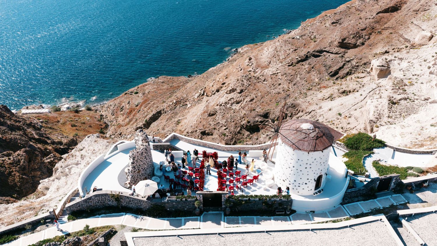 Aerial view of a white windmill wedding venue on rocky Santorini cliffside overlooking the deep blue Aegean Sea.