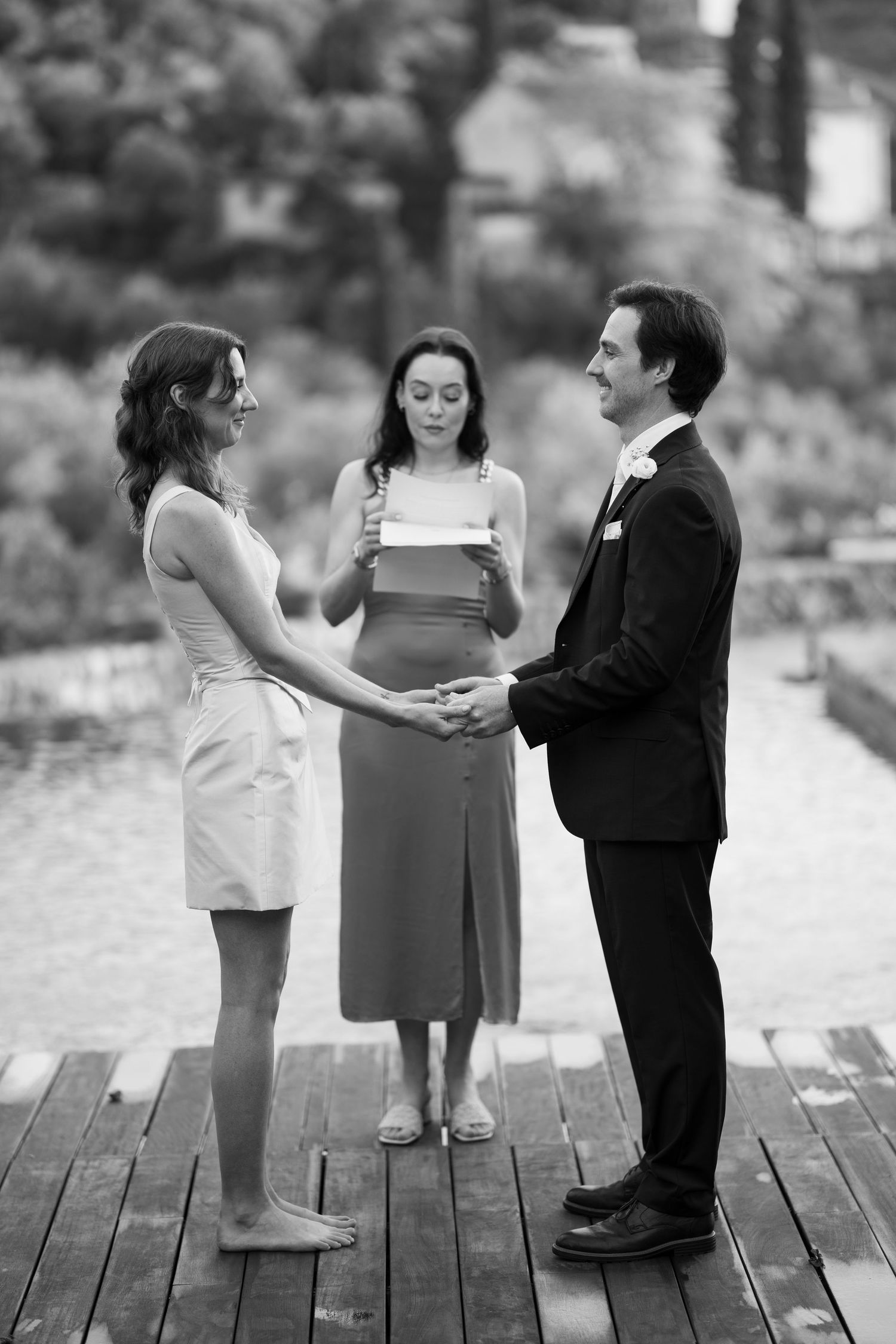 Barefoot bride and groom in a suit hold hands on a wooden deck as an officiant reads during an outdoor wedding ceremony.